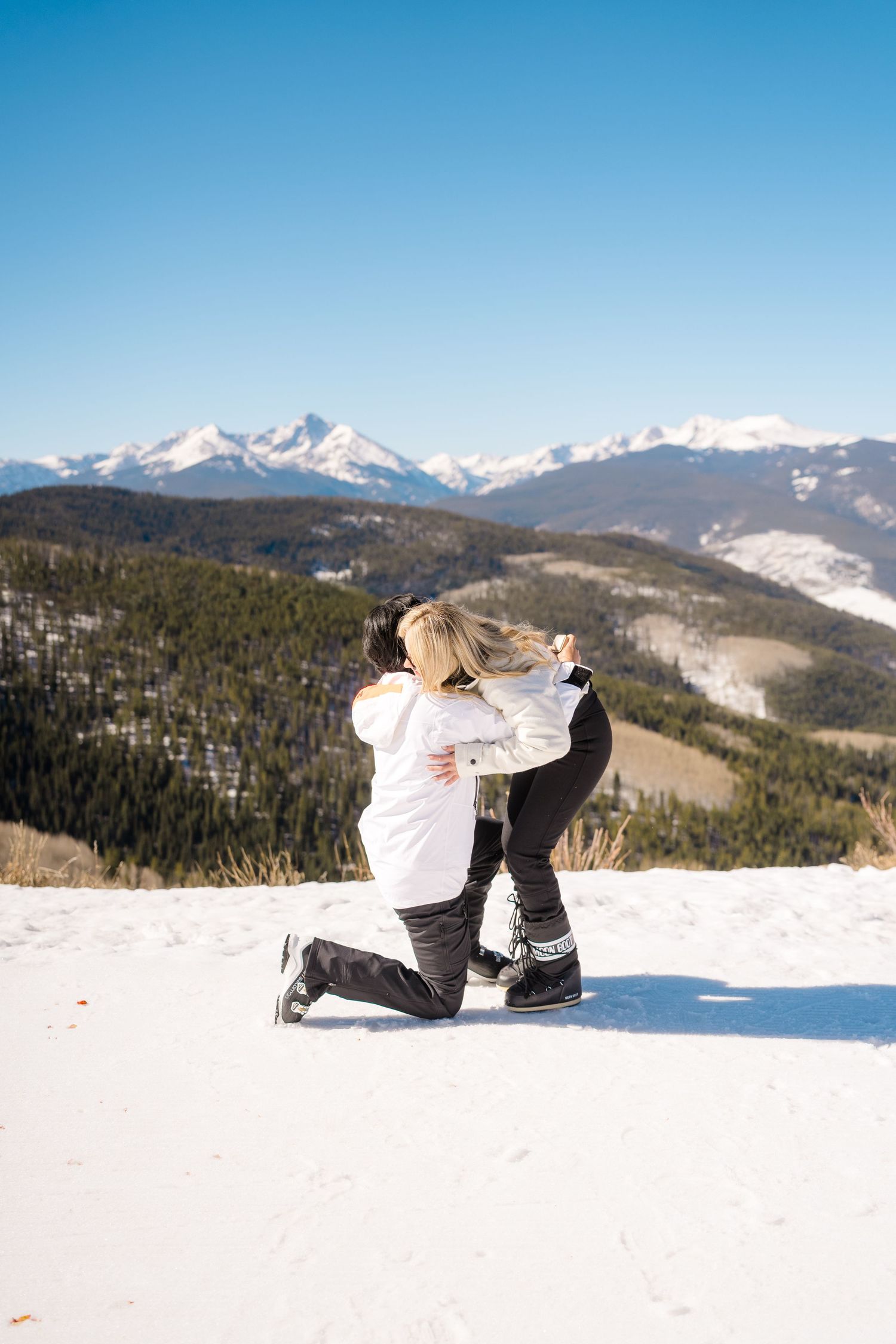 Couple embracing on snowy mountain top during a winter proposal with scenic snow-capped peaks in background.