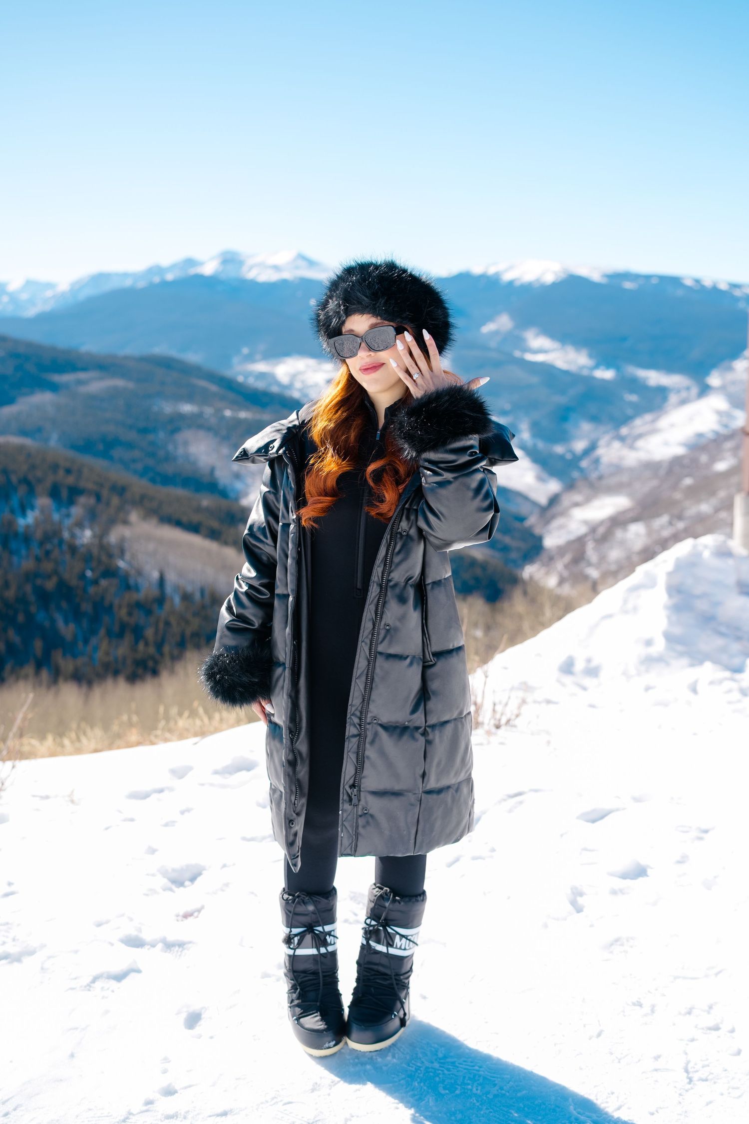 Person in a black puffer coat and winter boots poses on a snowy mountain slope with scenic mountain views in background.
