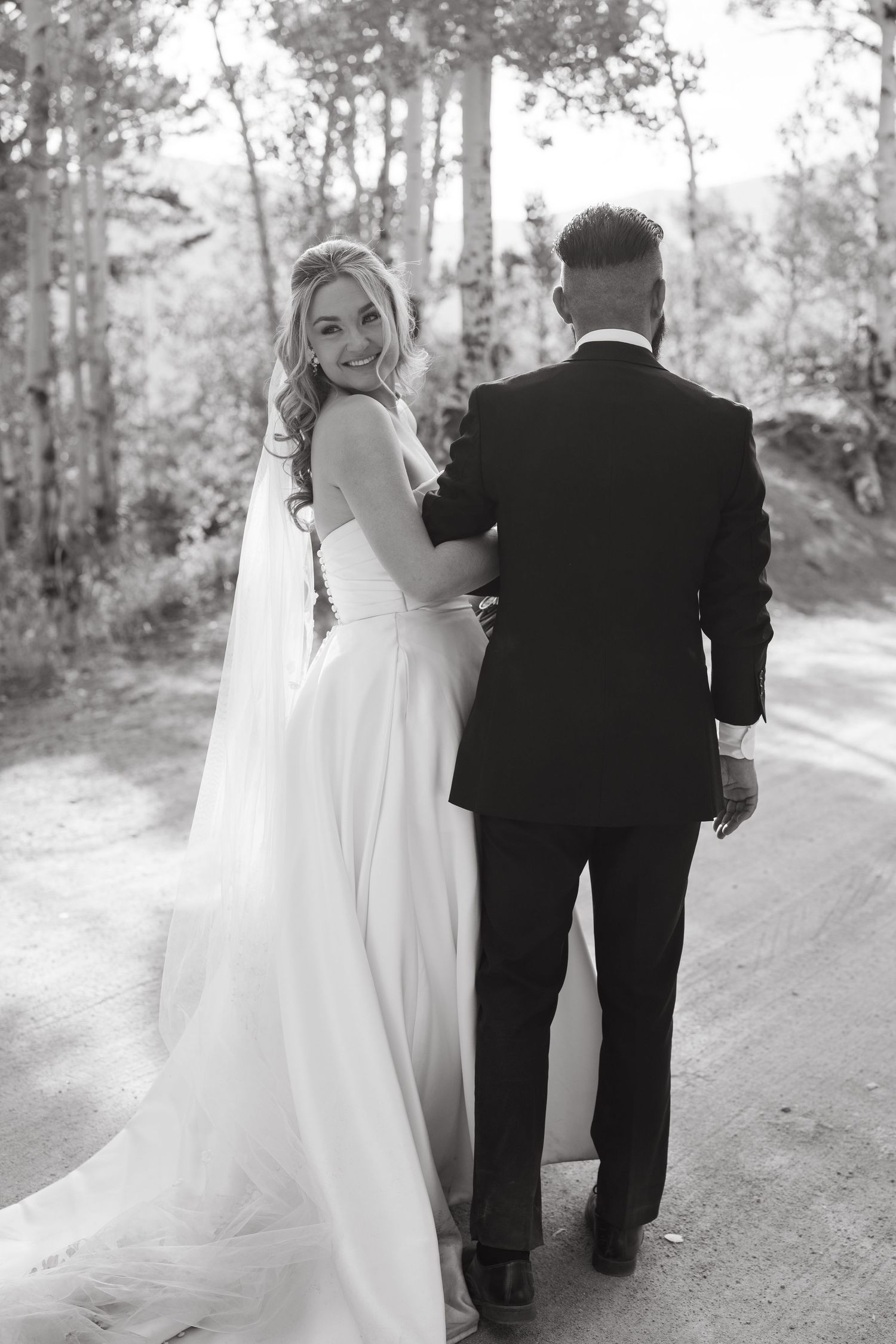 Black and white photo of newlyweds walking along forest path with bride looking back over shoulder.