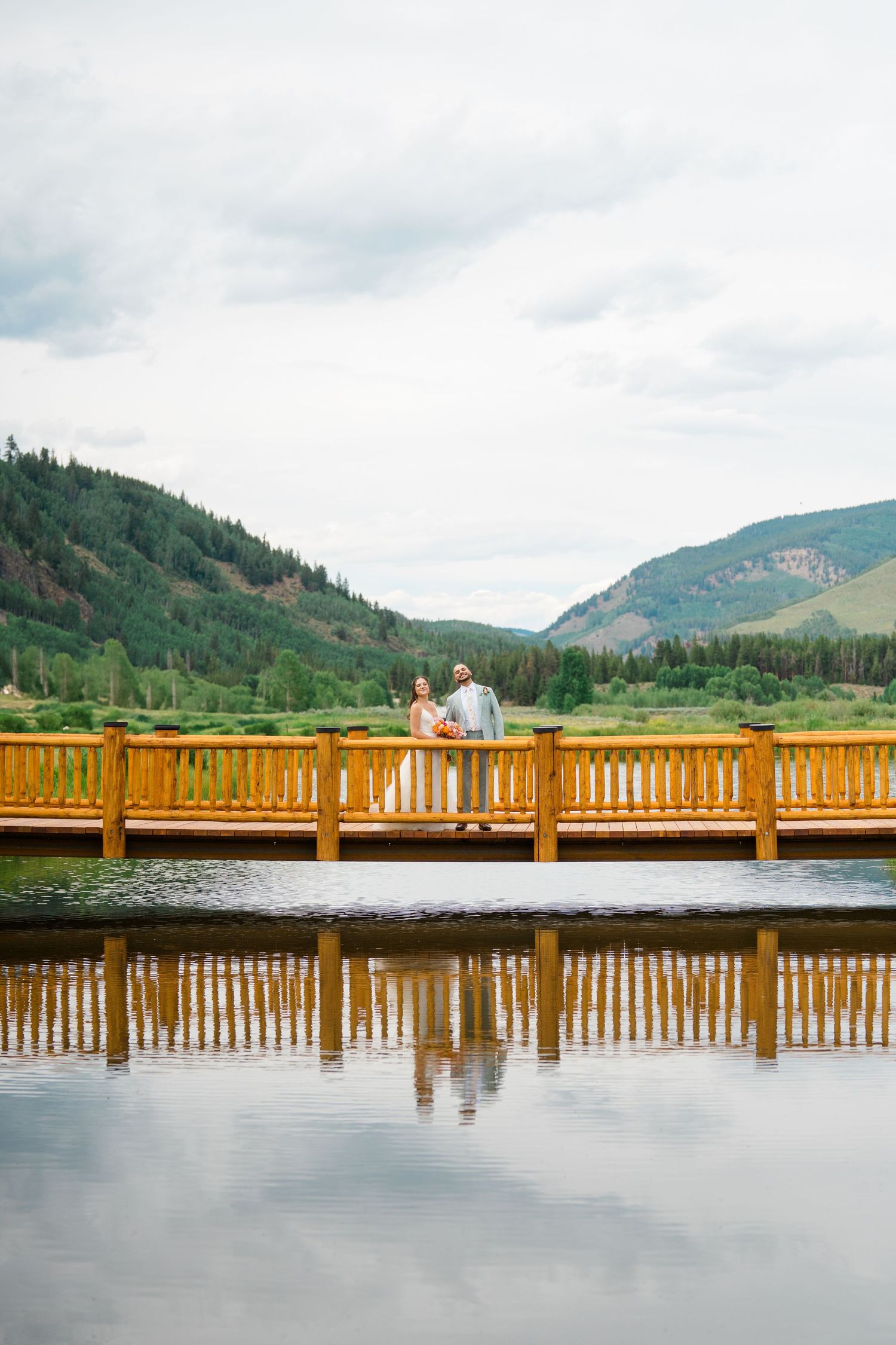 A wooden dock stretches across a serene lake with mountains and evergreen forests in the background under cloudy skies.