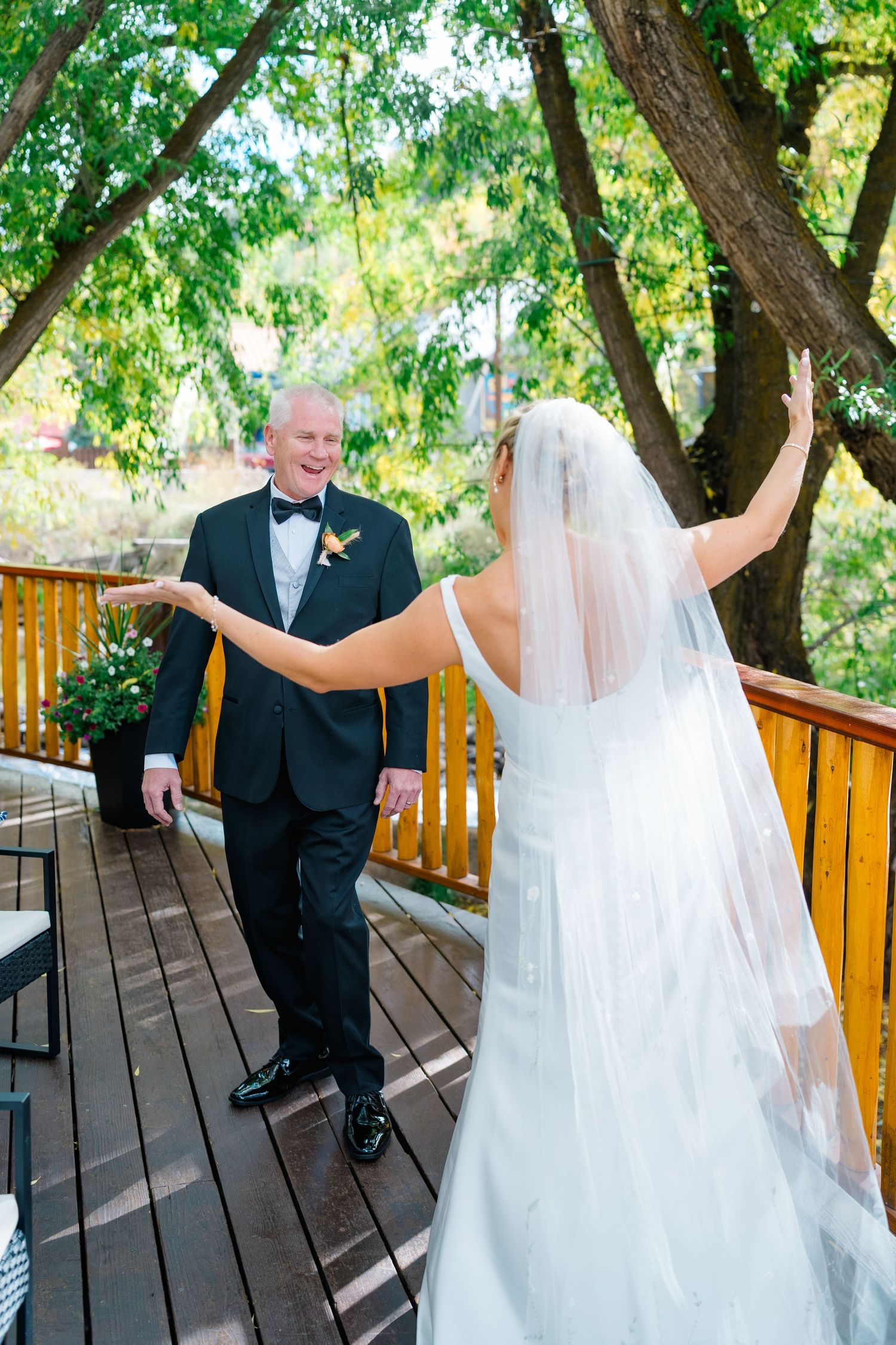 Wedding couple shares playful dance moves on wooden deck surrounded by green trees.