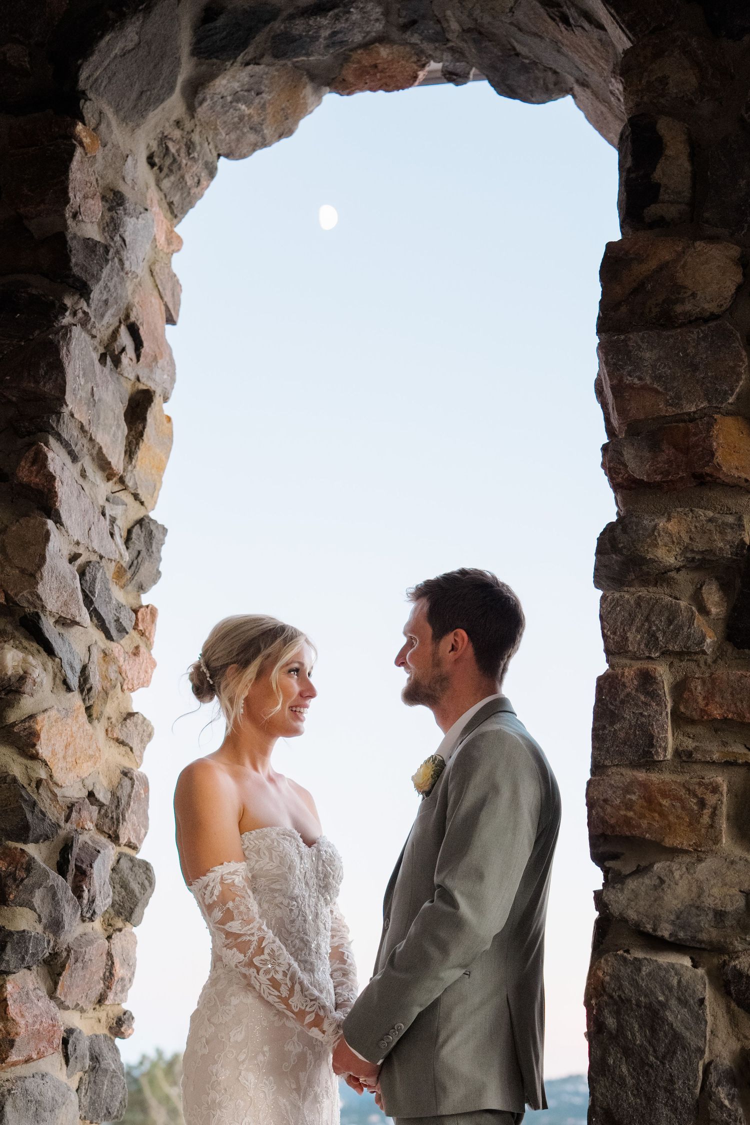 A romantic wedding couple shares a tender moment framed by a rustic stone archway at sunset.