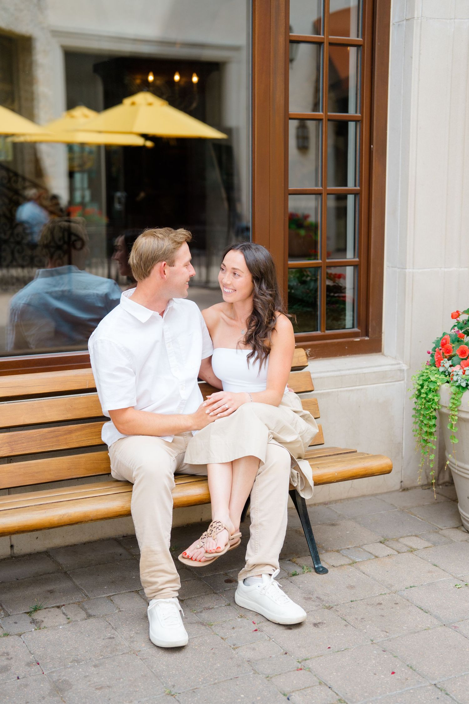 A couple dressed in white sits together on a wooden bench outside a restaurant with yellow umbrellas visible through windows.