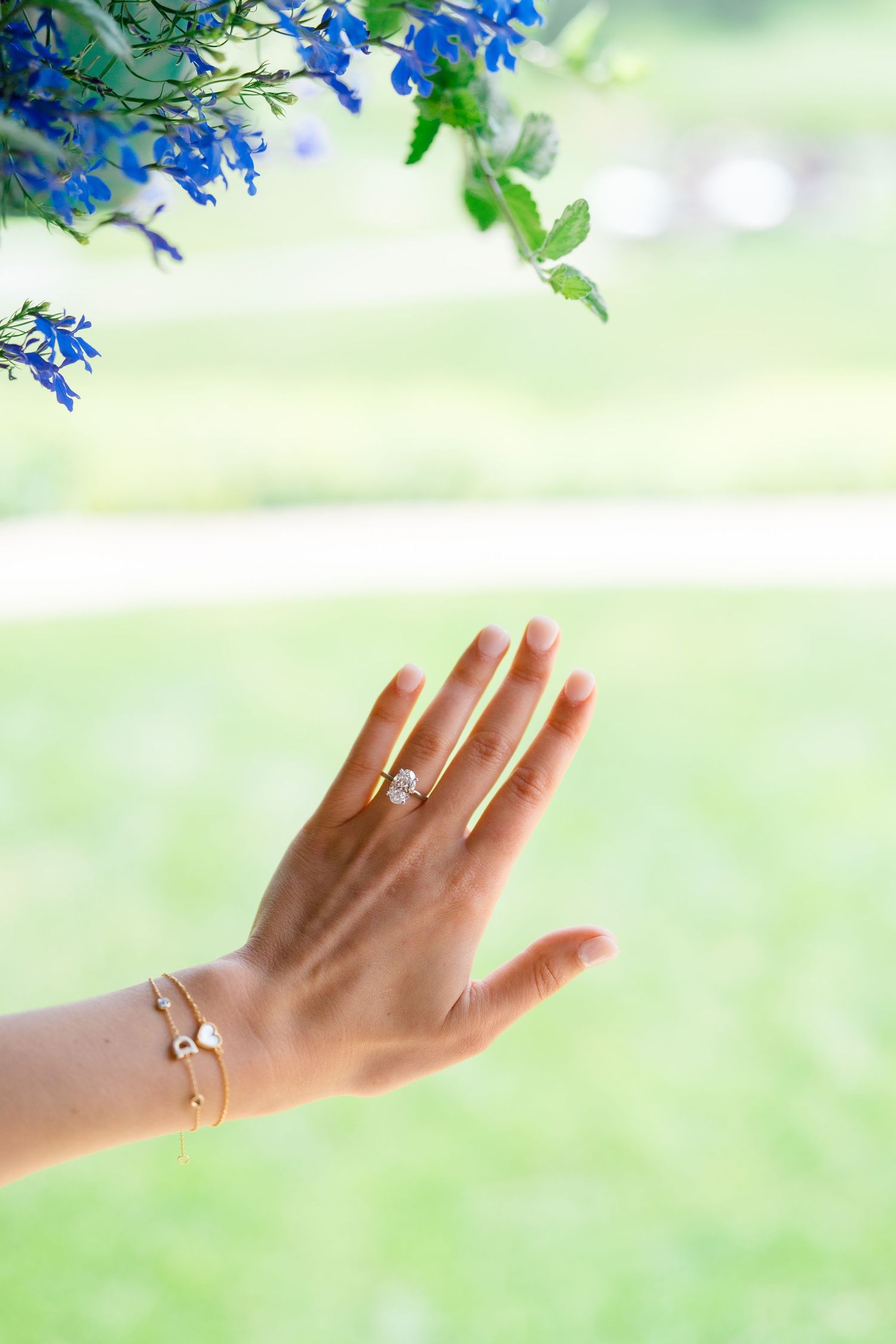 Delicate hand with engagement ring extended against soft green background with blue flowers above.