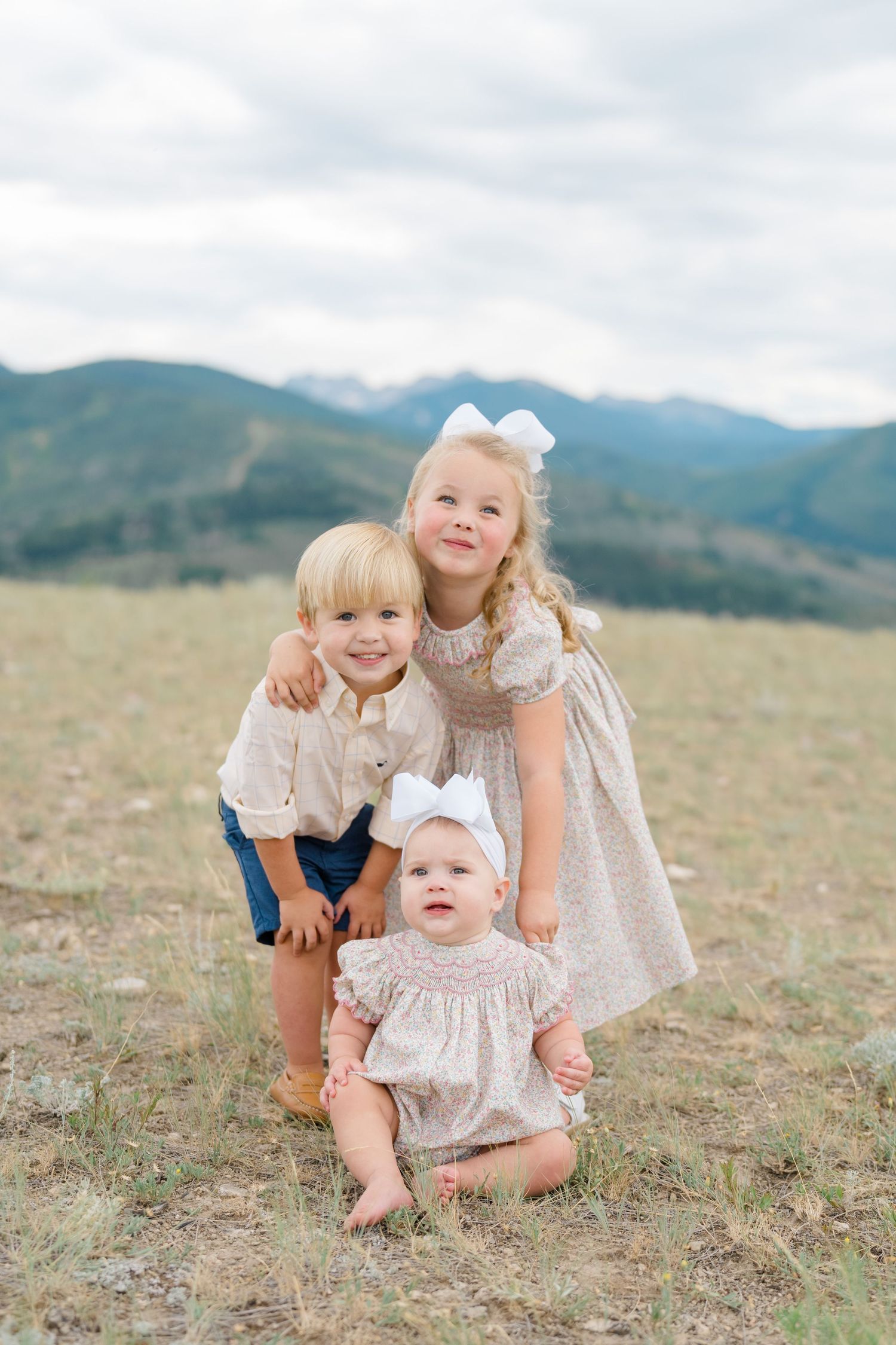 Three young children play together on a grassy hillside with mountains in the background.