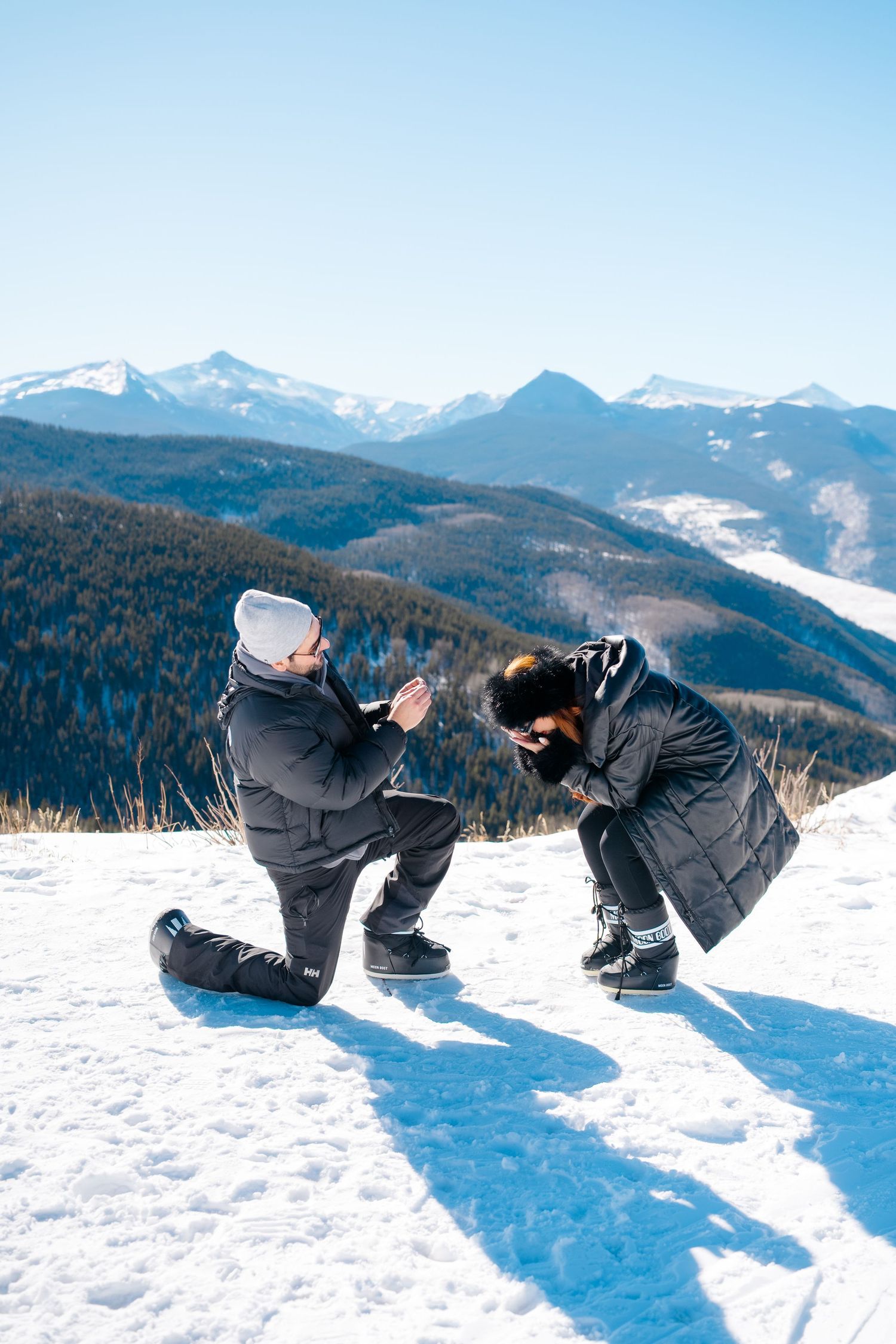 Two people in winter gear crouch down to take photos of the snowy mountain landscape on a bright sunny day at vail mountaintop proposal.