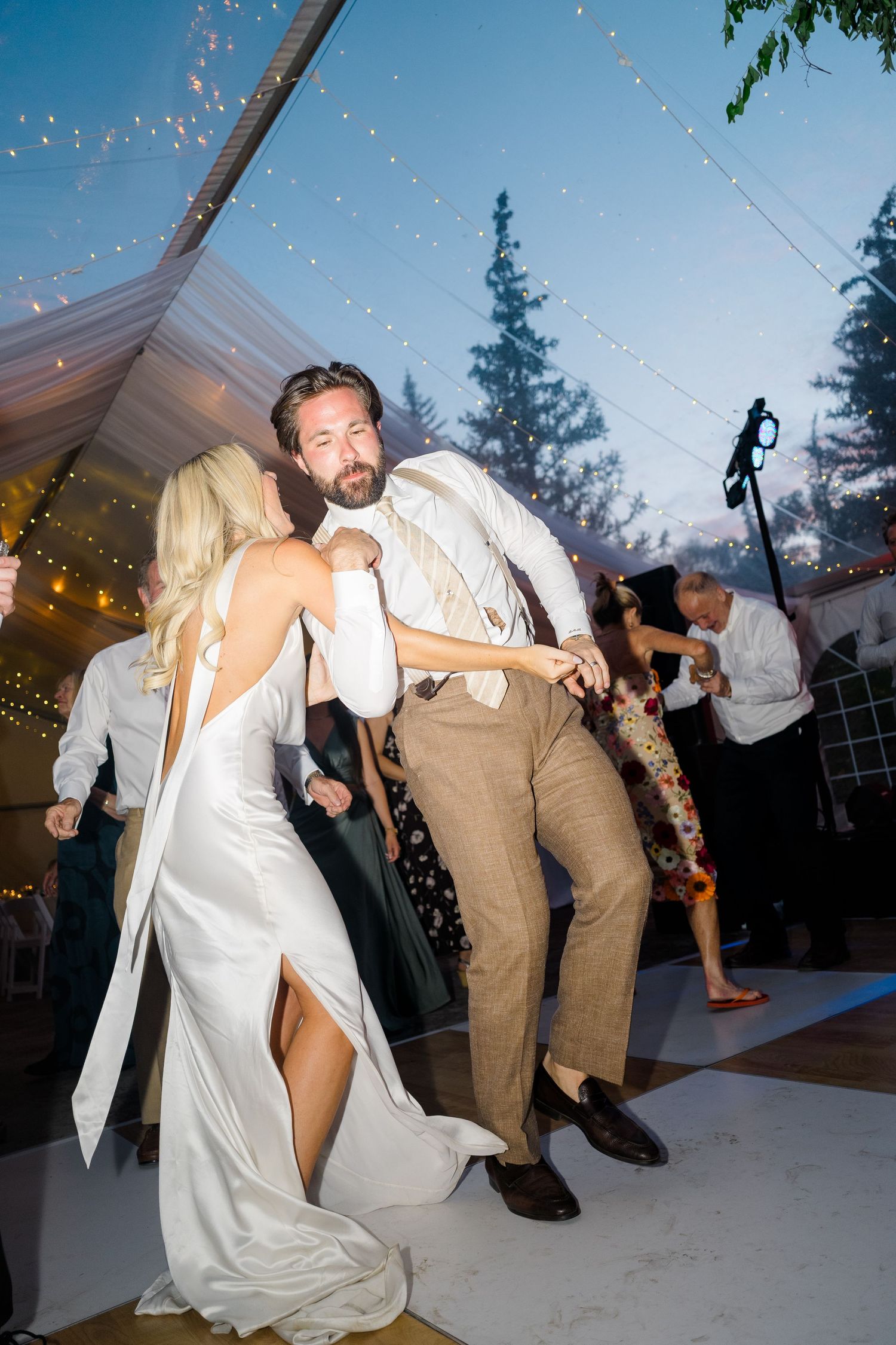 Bride and groom dancing energetically at outdoor wedding reception under string lights at dusk.