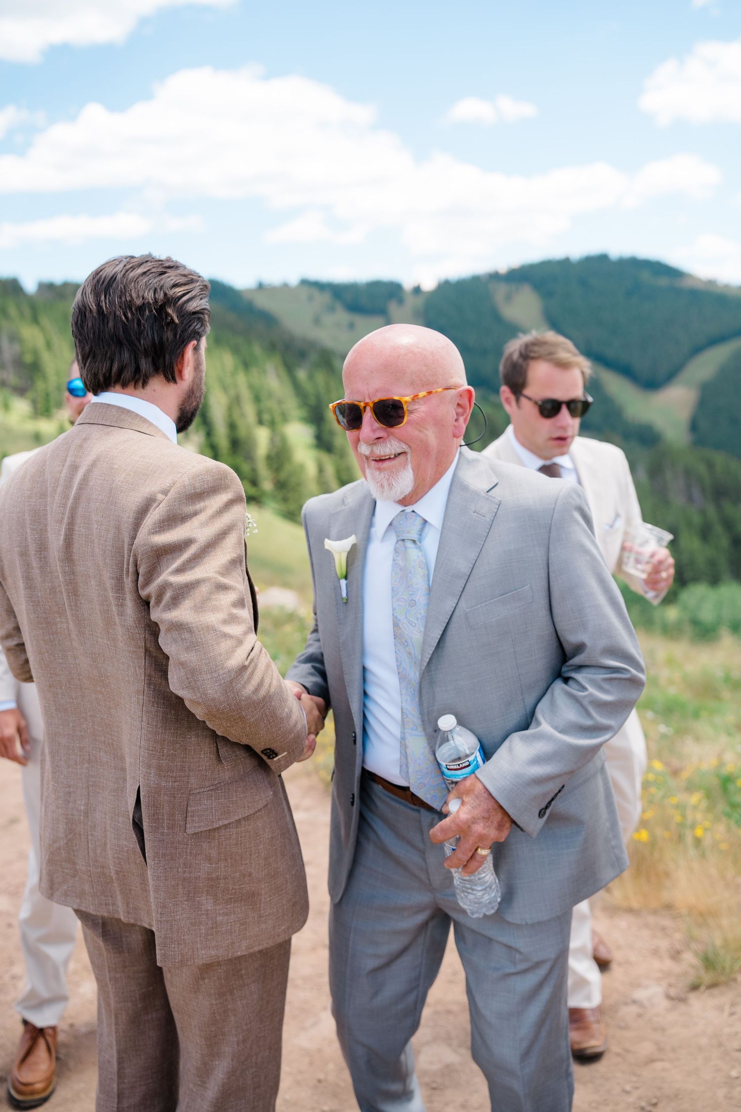 Elderly man in gray suit and orange sunglasses shaking hands at an outdoor mountain wedding ceremony.