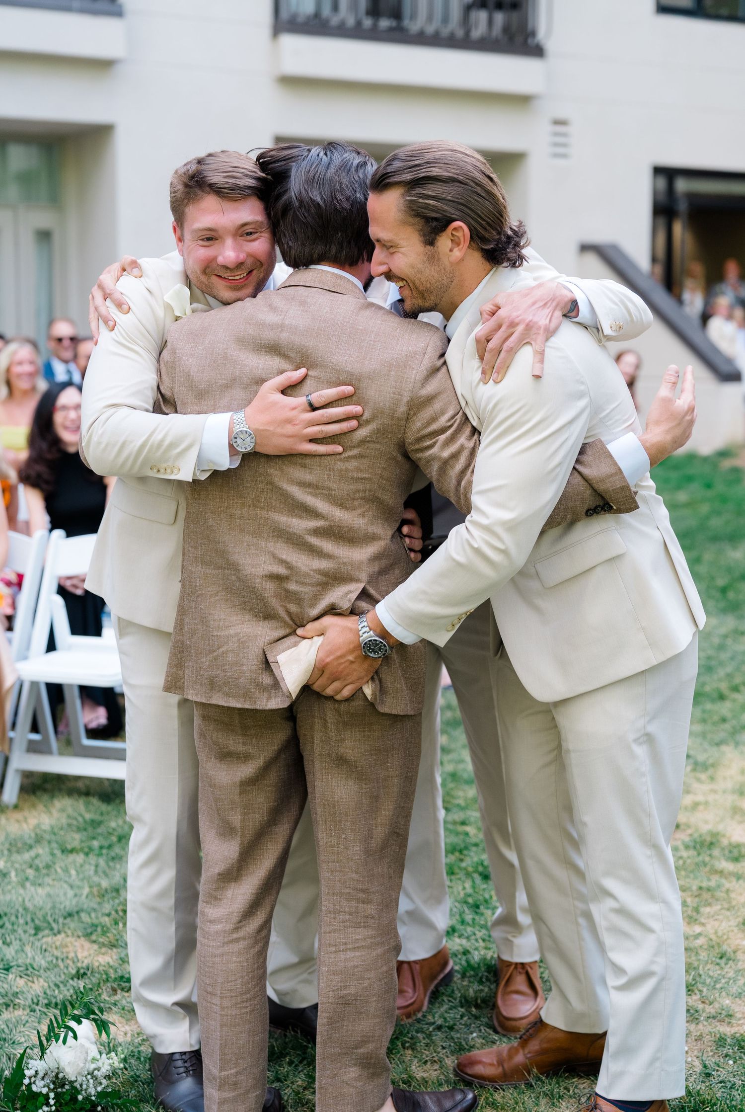 Three men in suits sharing a group hug at an outdoor wedding ceremony on a sunny day.