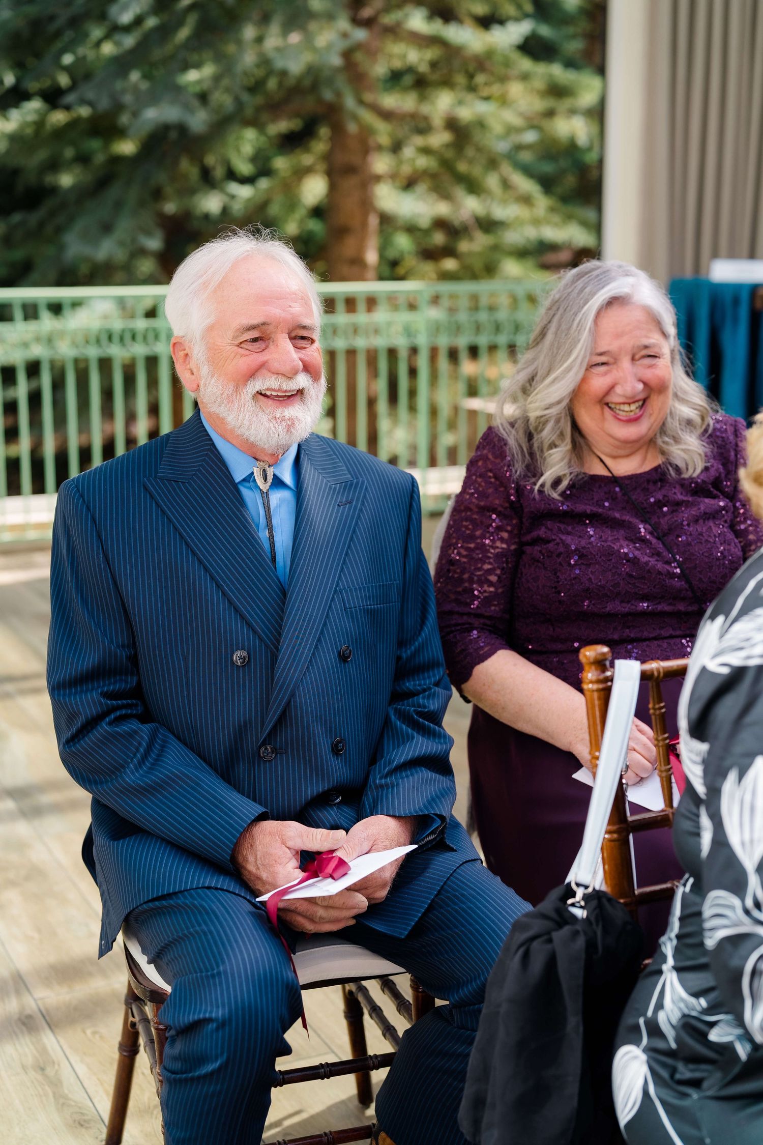 Elderly couple smiling and laughing at a wedding ceremony, man in navy suit, woman in purple sequin dress.
