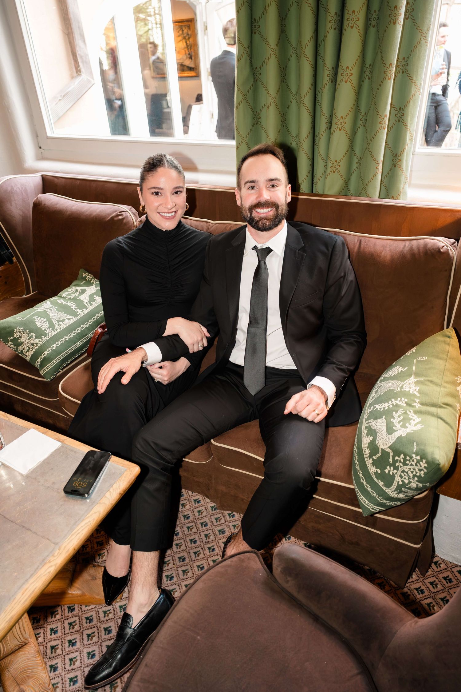 A smiling couple dressed in black sit together on a brown leather sofa with green decorative pillows.