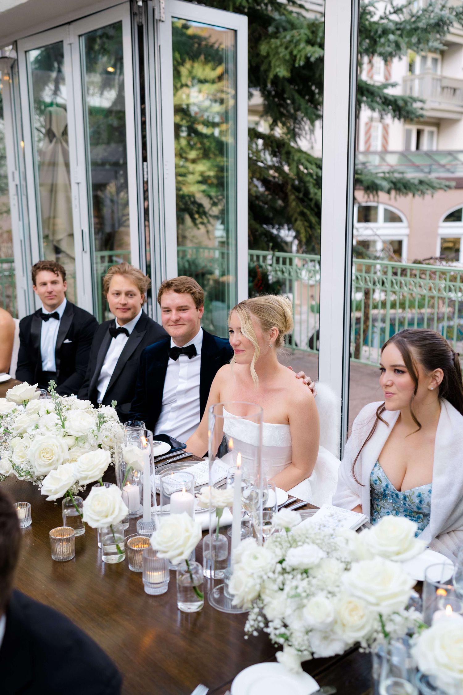 Elegant wedding reception guests seated at a decorated table with white roses and candles in a glass-walled venue.
