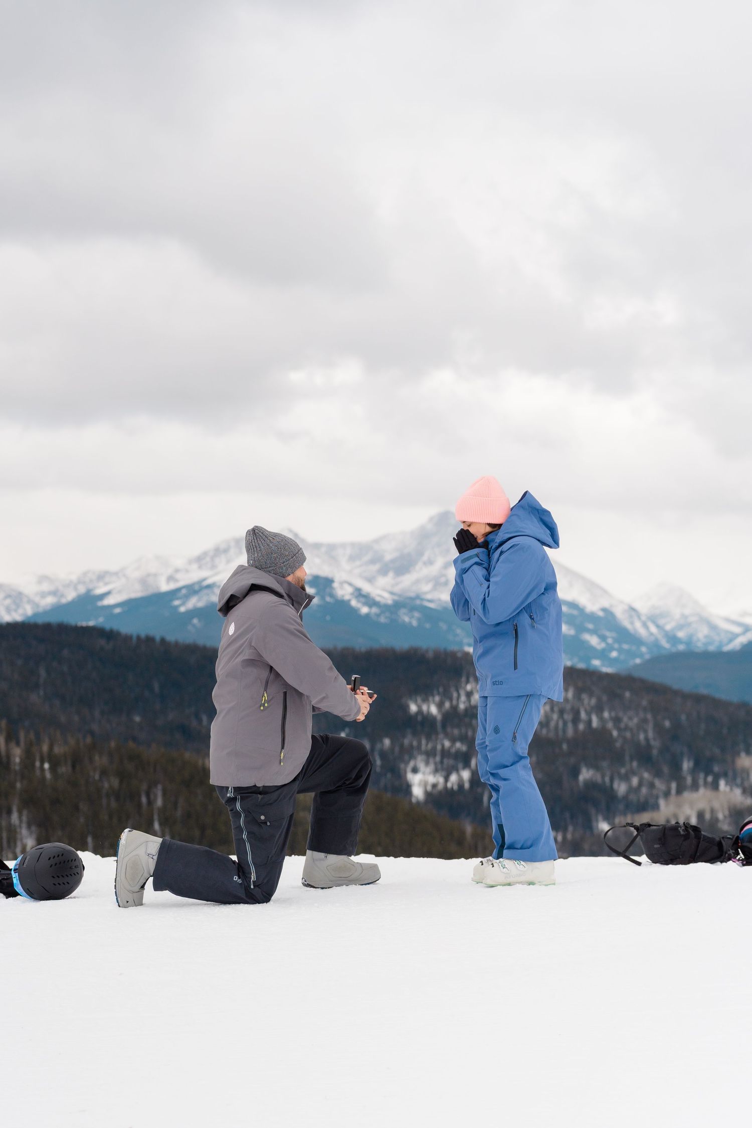 A winter proposal scene with two people in snow gear against snowy mountain peaks and cloudy skies.