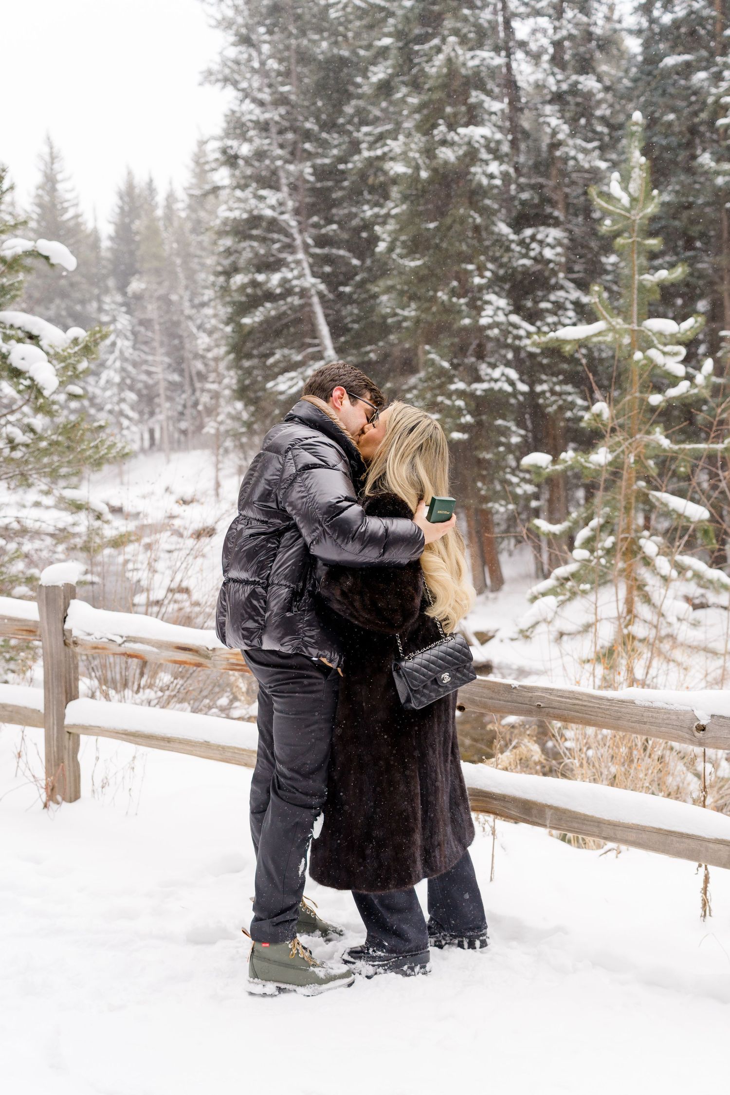 A couple shares a romantic kiss in a snowy winter landscape with evergreen trees and a wooden fence in the background.