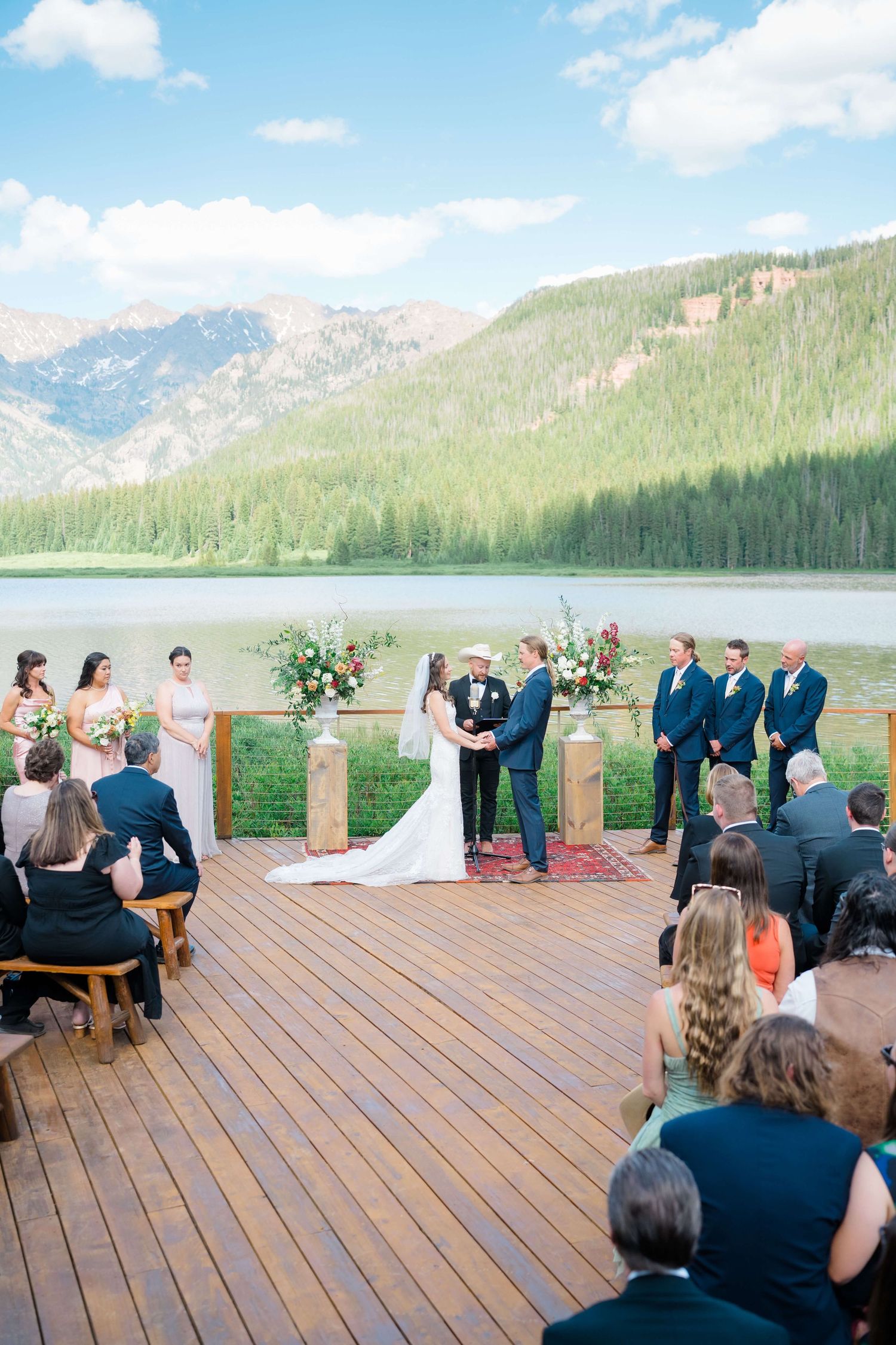 Outdoor mountain Piney River Ranch wedding ceremony on wooden deck with bride, groom, wedding party, and guests under blue sky.