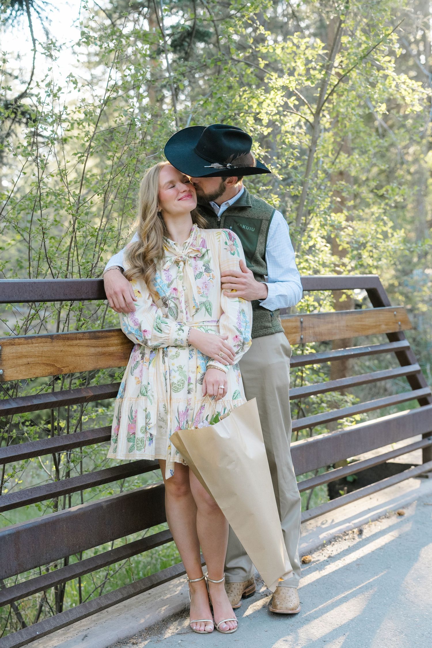 Couple sharing a romantic kiss on a wooden bridge, woman in floral dress holding wrapped flowers, man in cowboy hat.