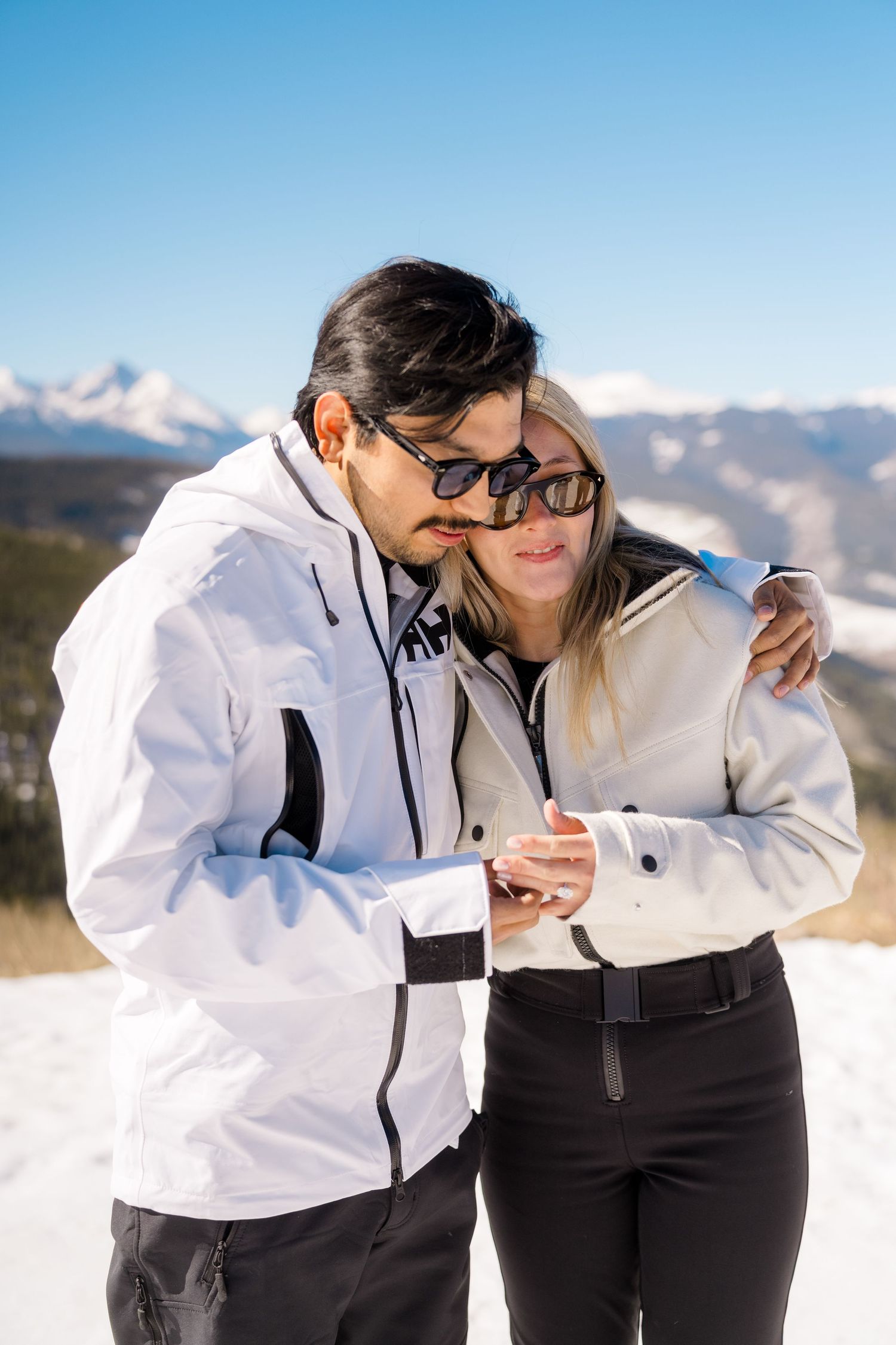 Couple in white ski jackets embracing on snowy mountain slope with blue sky and Alpine peaks behind them.