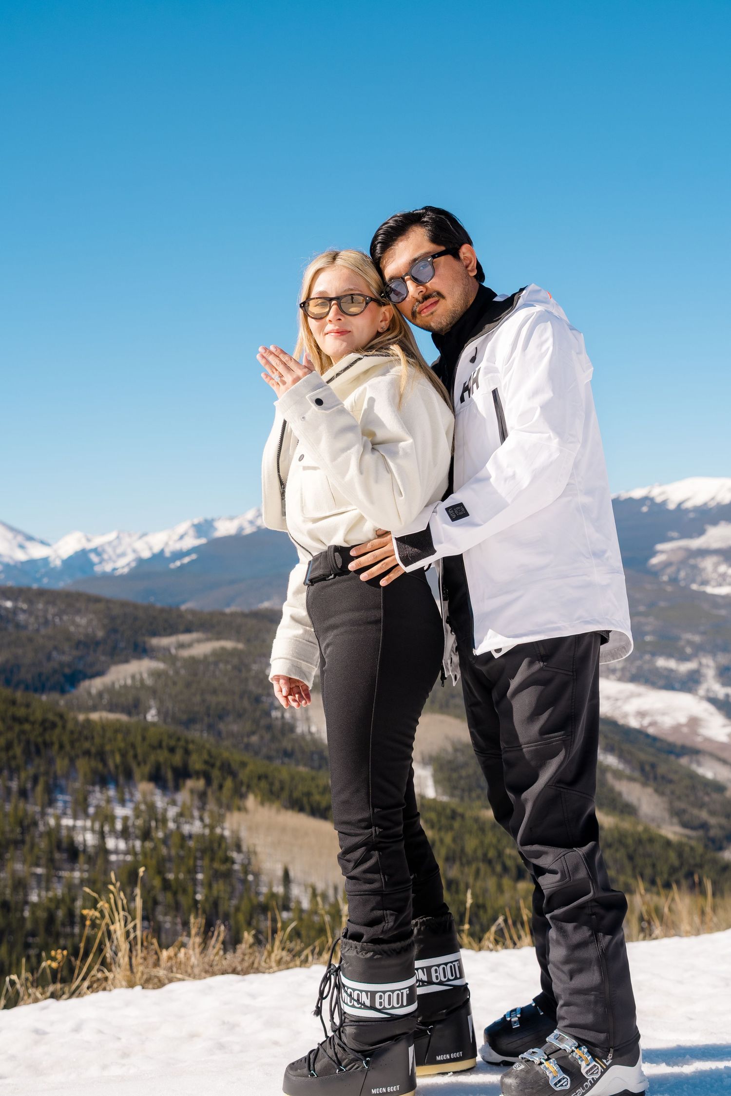 Couple in white ski jackets posing on snowy mountain with blue sky and pine trees in background.