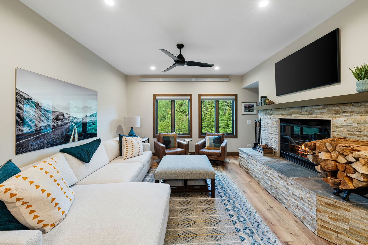 Modern living room with sectional sofa, stone fireplace, ceiling fan, wall-mounted TV, and large windows with forest views.