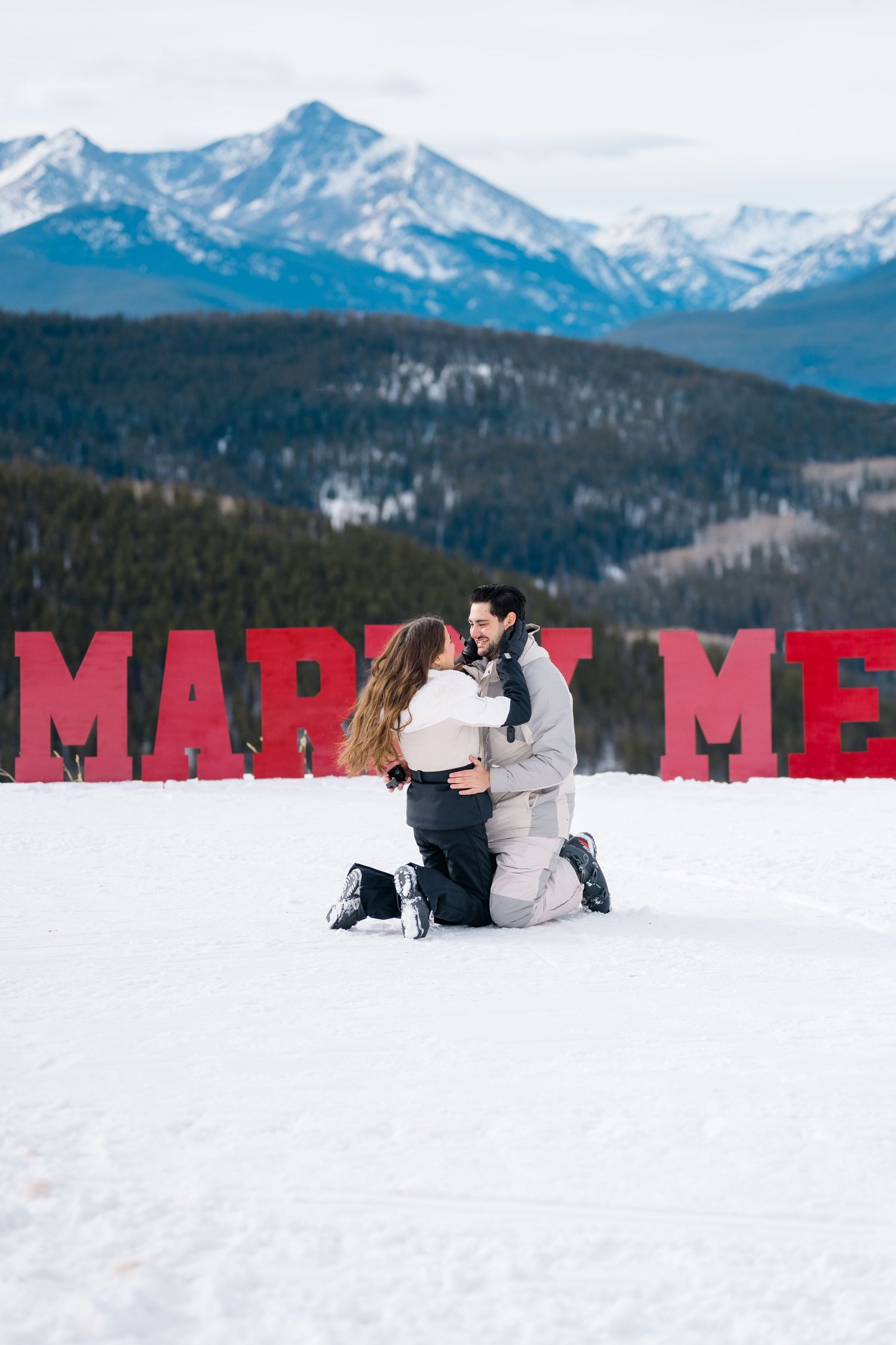 Two people sit together in snow with snowy mountain peaks and large red letters visible in winter landscape.