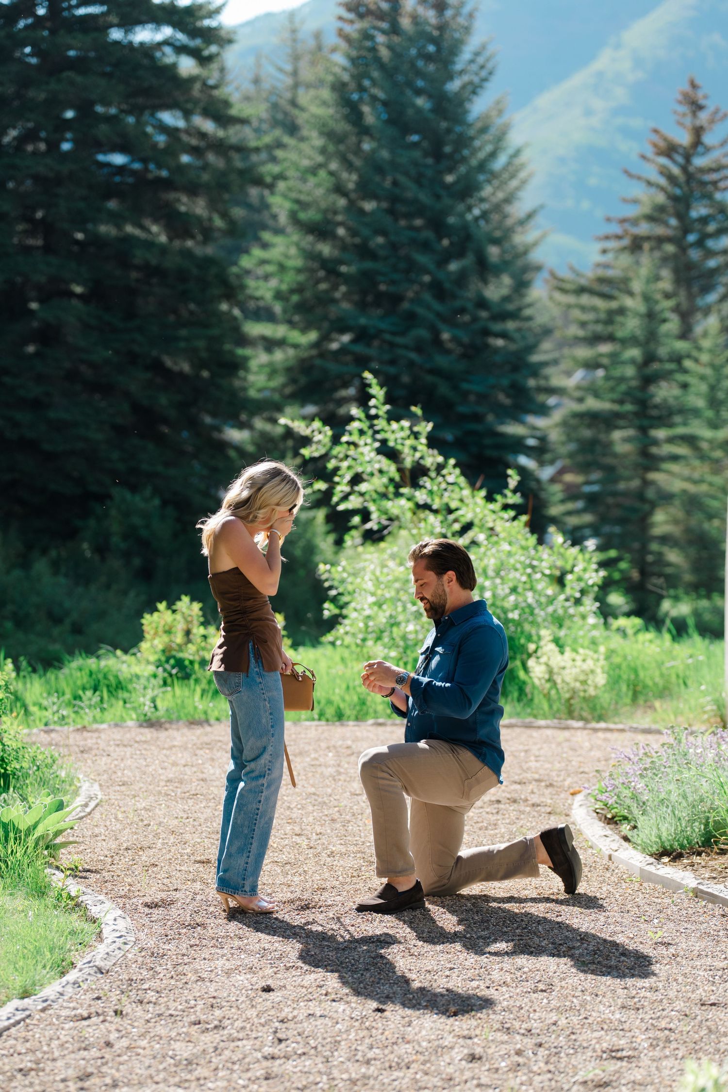 A couple shares a special moment on a mountain trail surrounded by evergreen trees and mountain views.