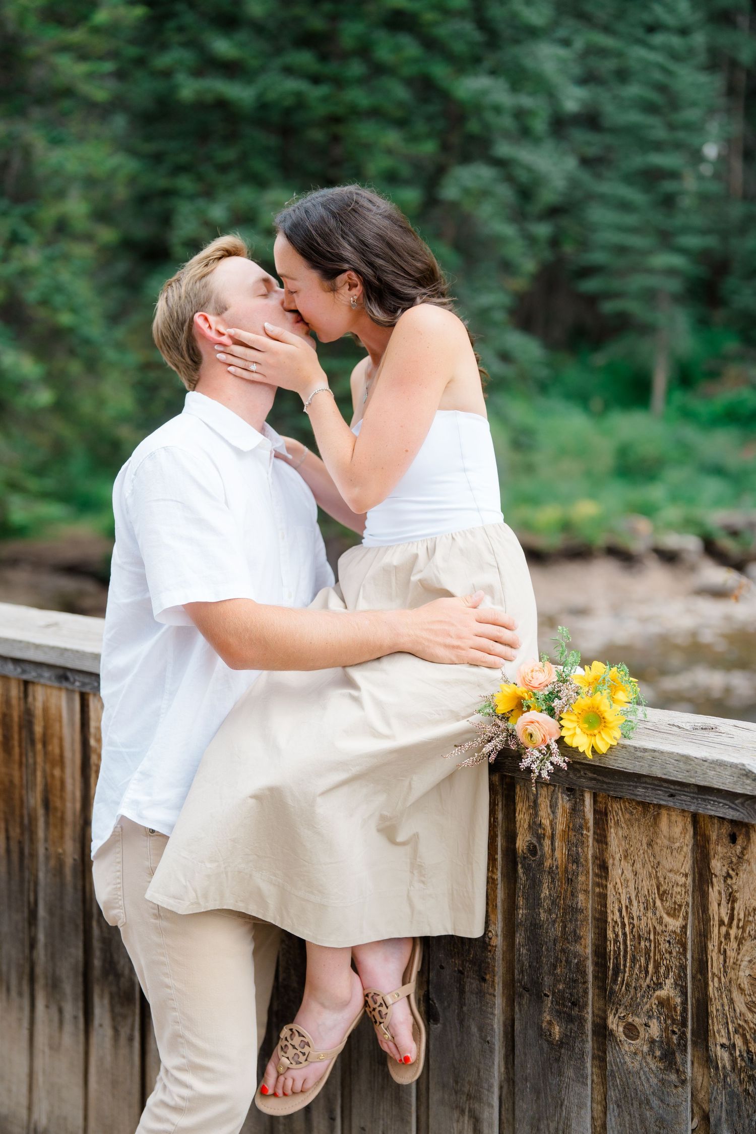 A romantic couple shares tender kisses while sitting on a wooden railing by a lake with sunflowers nearby.
