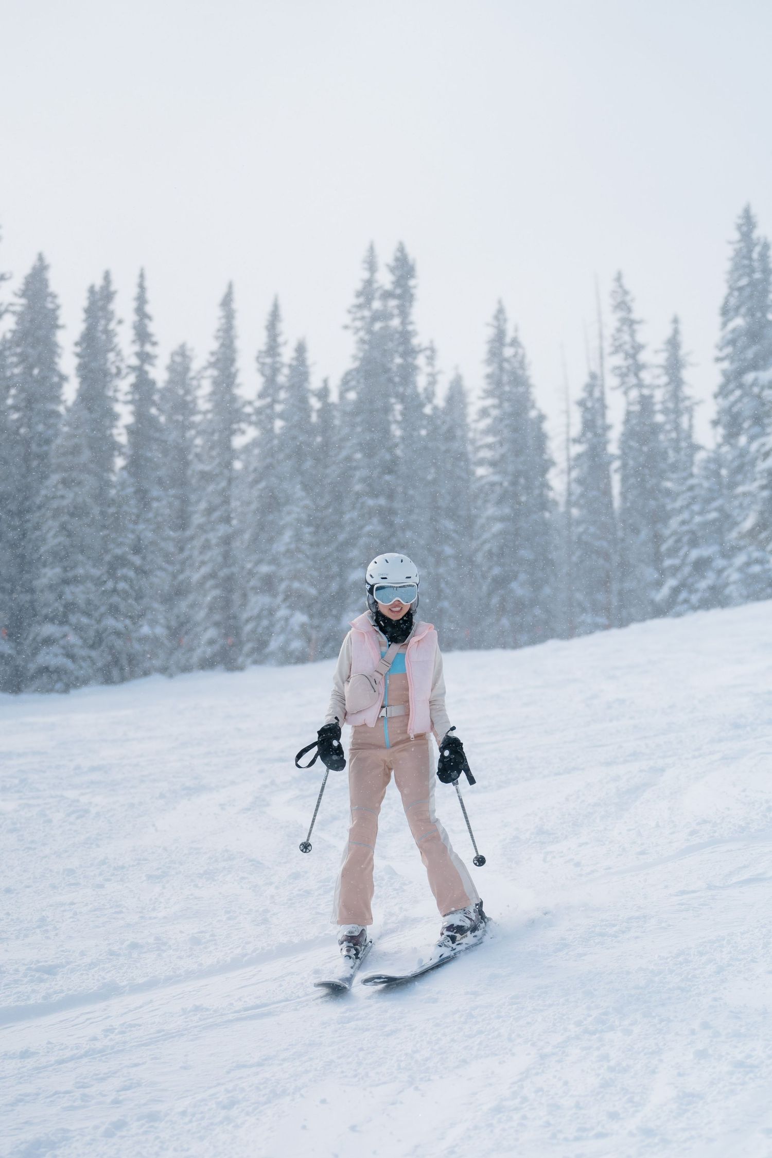 A skier in light pink ski gear glides down a snowy slope surrounded by snow-covered evergreen trees on a winter day.