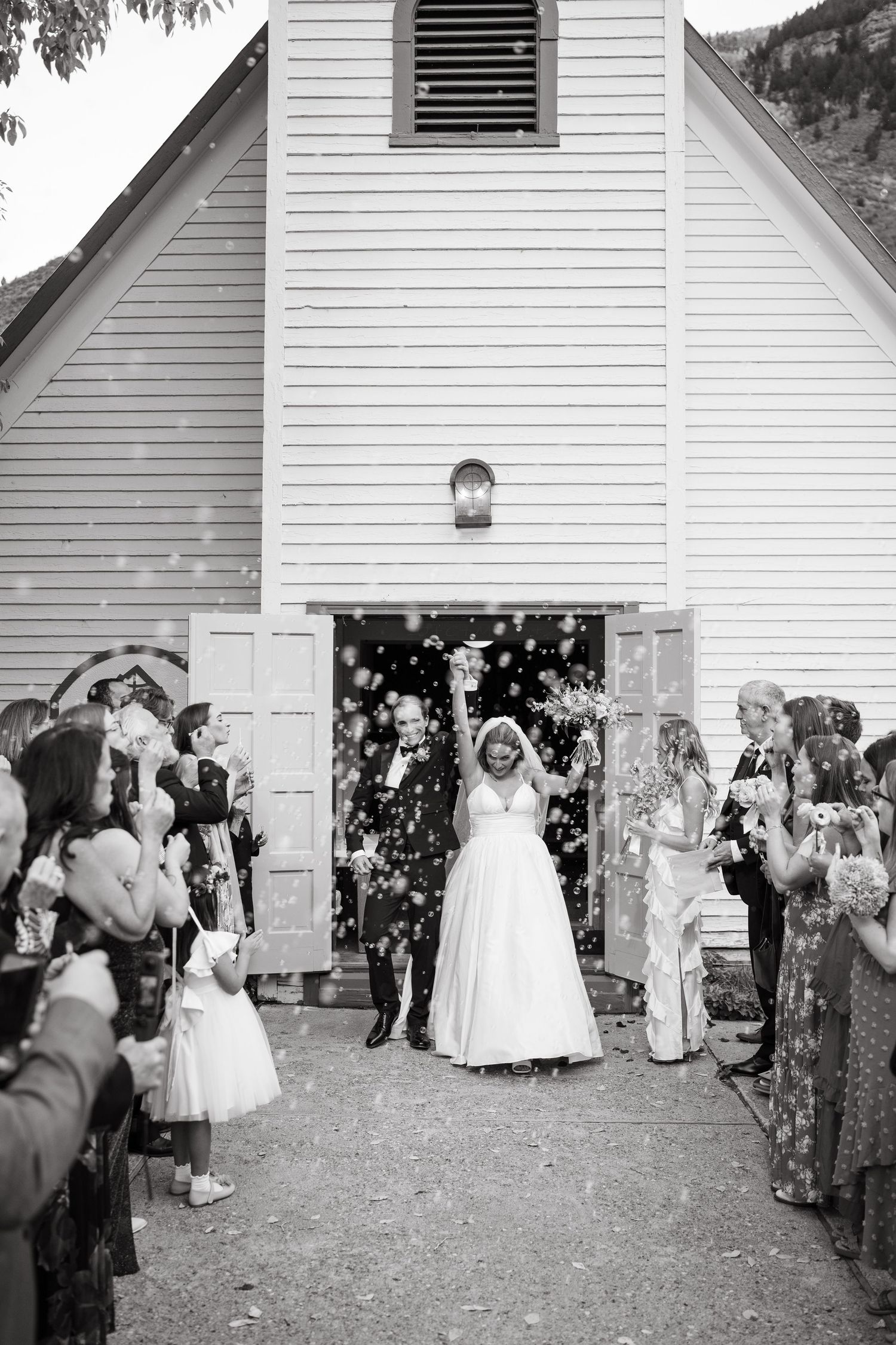 Wedding guests throw flower petals outside a white church after ceremony.