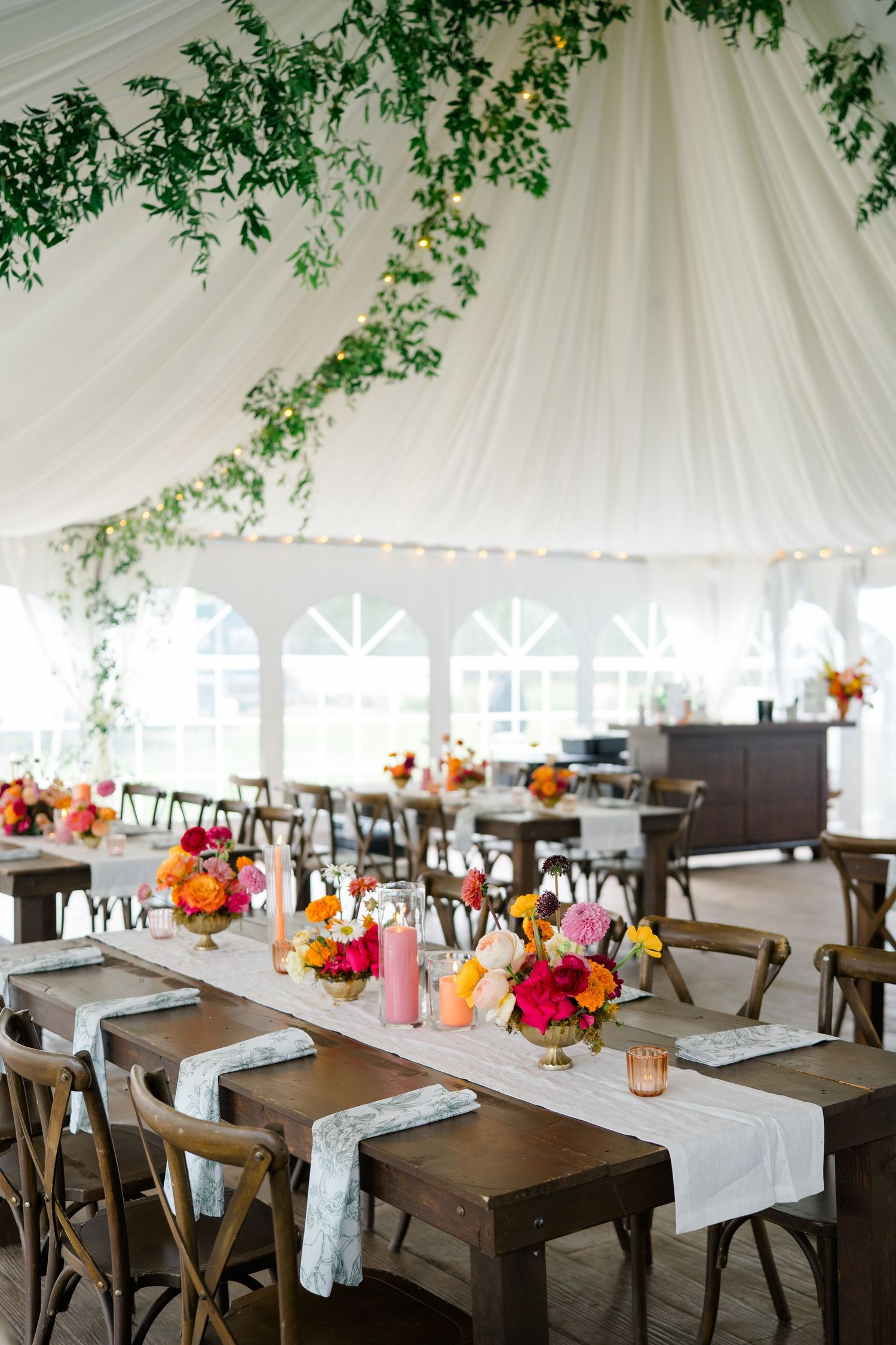 Rustic wedding reception setup with wooden tables and chairs under a white tent decorated with hanging greenery.