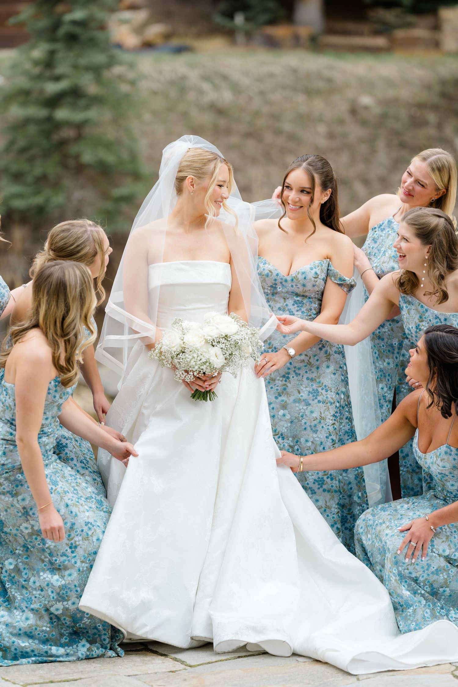 Bride in white gown holds bouquet surrounded by bridesmaids in blue floral dresses adjusting her veil outdoors.