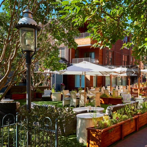 Outdoor patio dining area with white umbrellas, wicker chairs, and lush green trees beside a brick building.