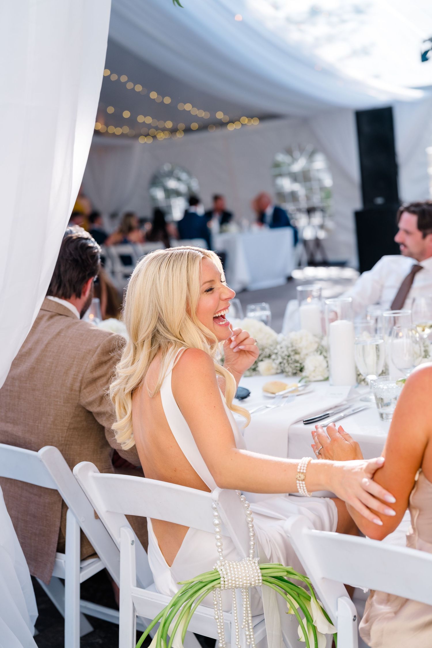 Blonde woman in white dress laughing joyfully at an elegant outdoor wedding reception under a white tent.