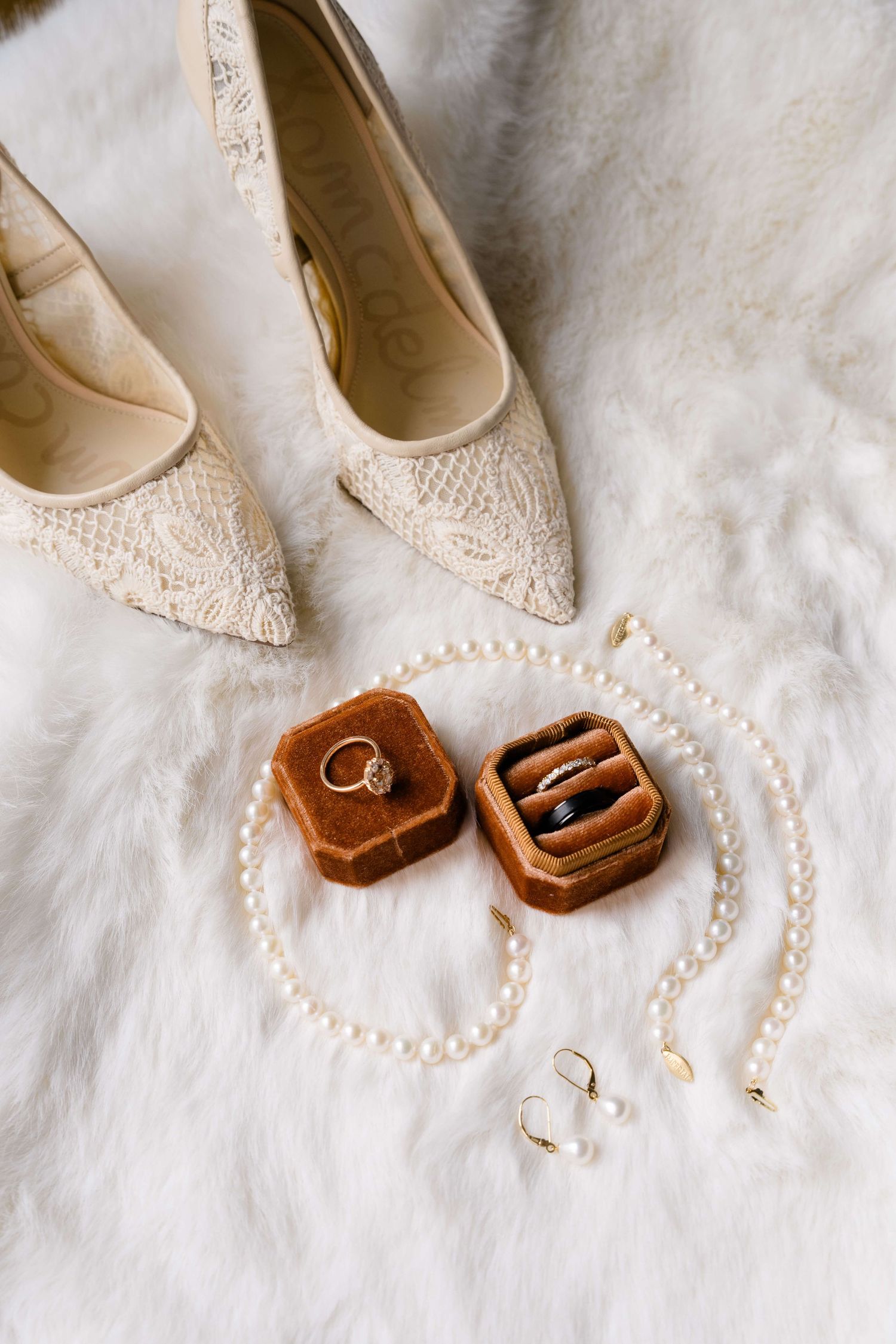 Bridal flat lay with lace heels, velvet ring box with wedding bands, pearl necklace, and gold earrings on white fur.