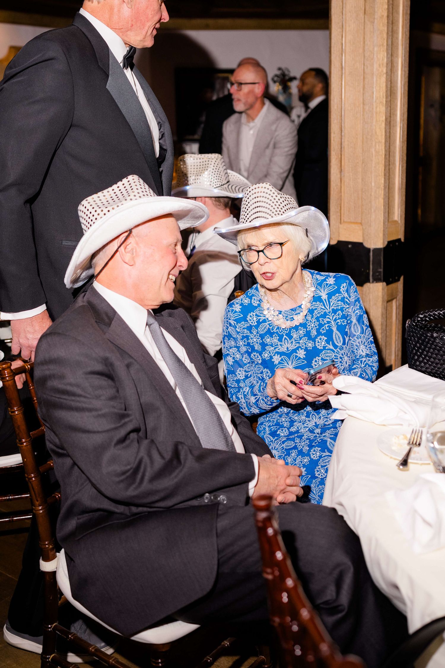 Elderly couple in white cowboy hats at a formal event, woman in blue floral dress with pearls, man in dark suit.