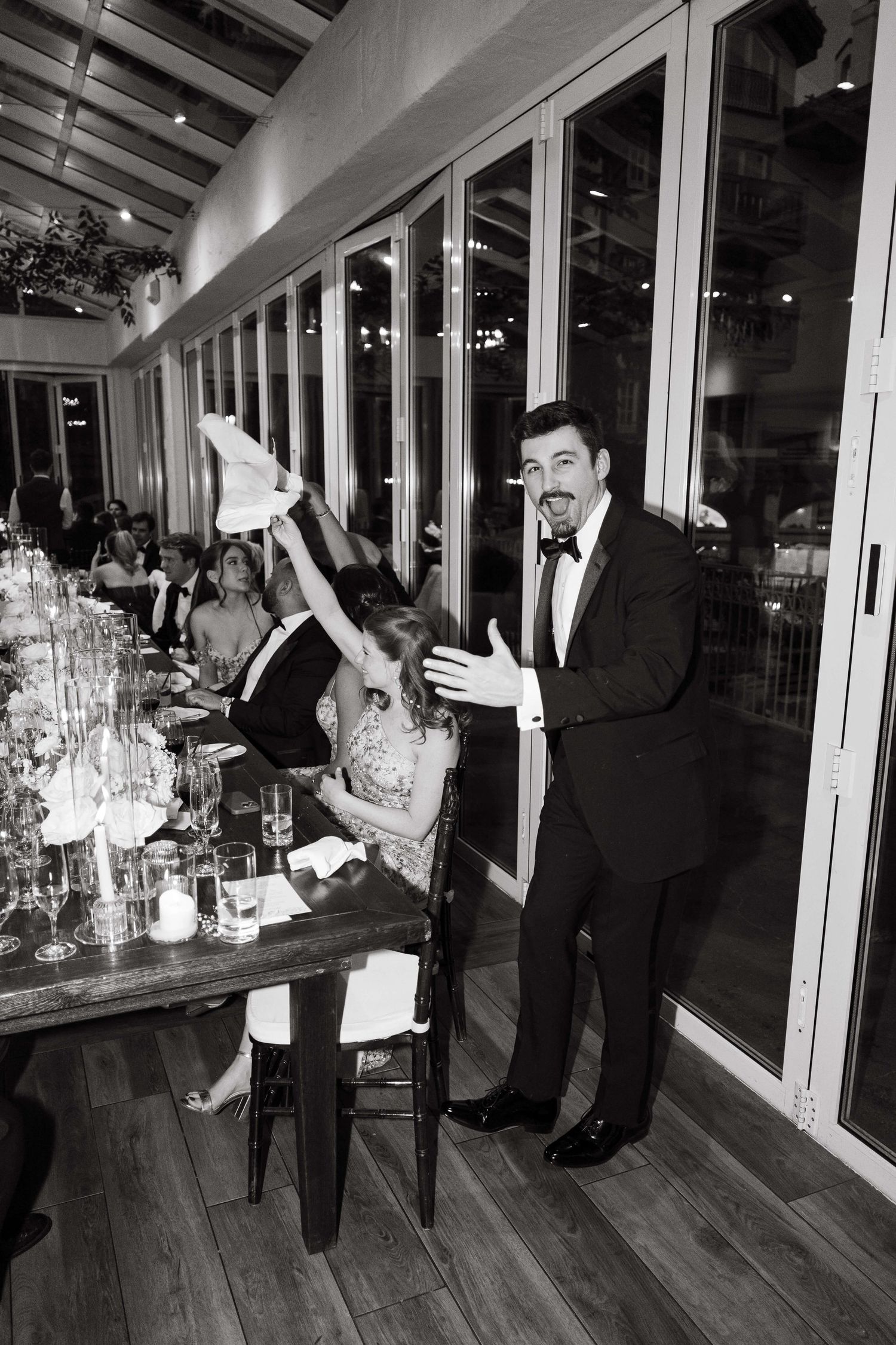 Black and white photo of a man in a tuxedo gesturing at a wedding reception dinner table with guests celebrating.