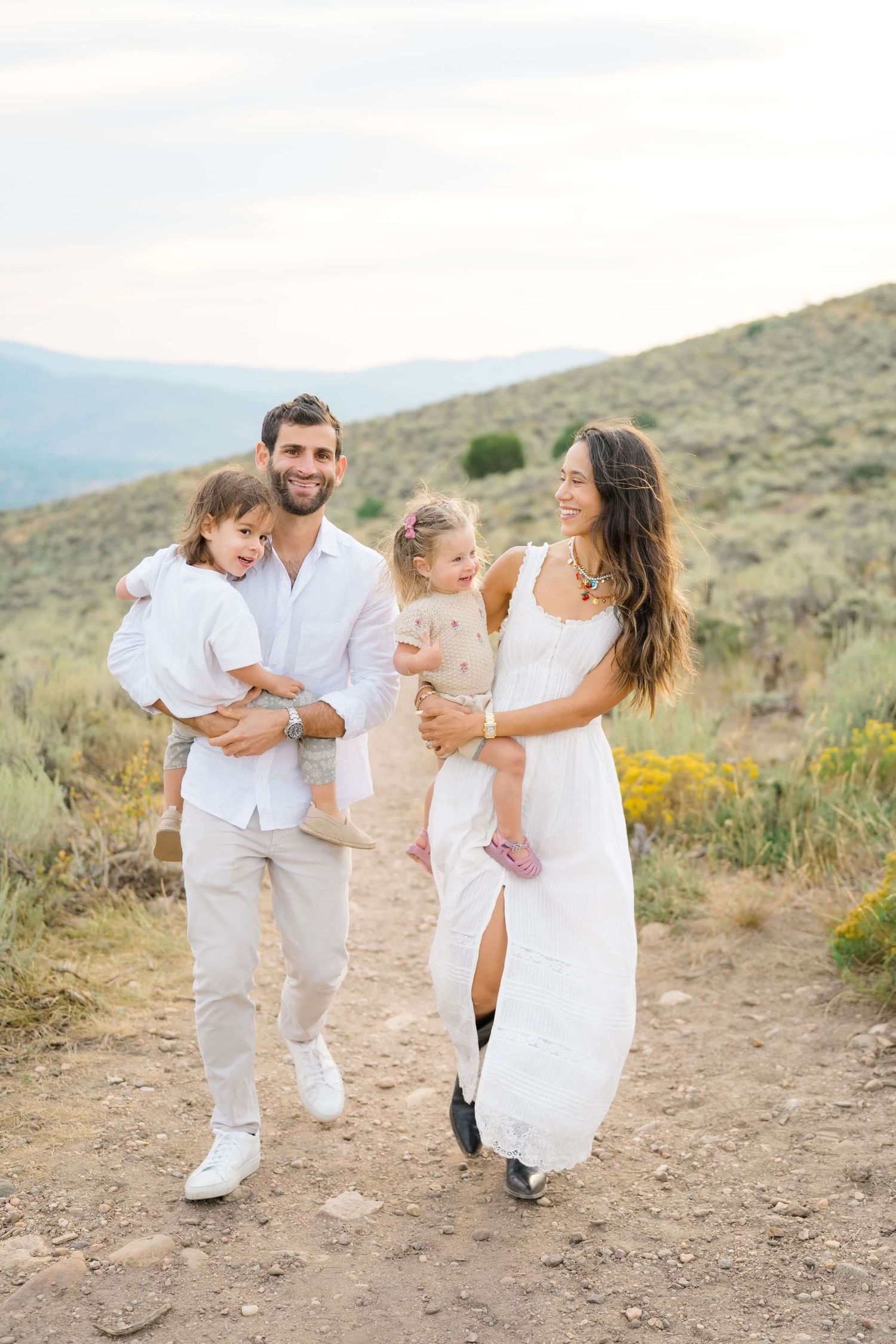 A family wearing white outfits walks together on a mountain trail during sunset in a desert landscape.