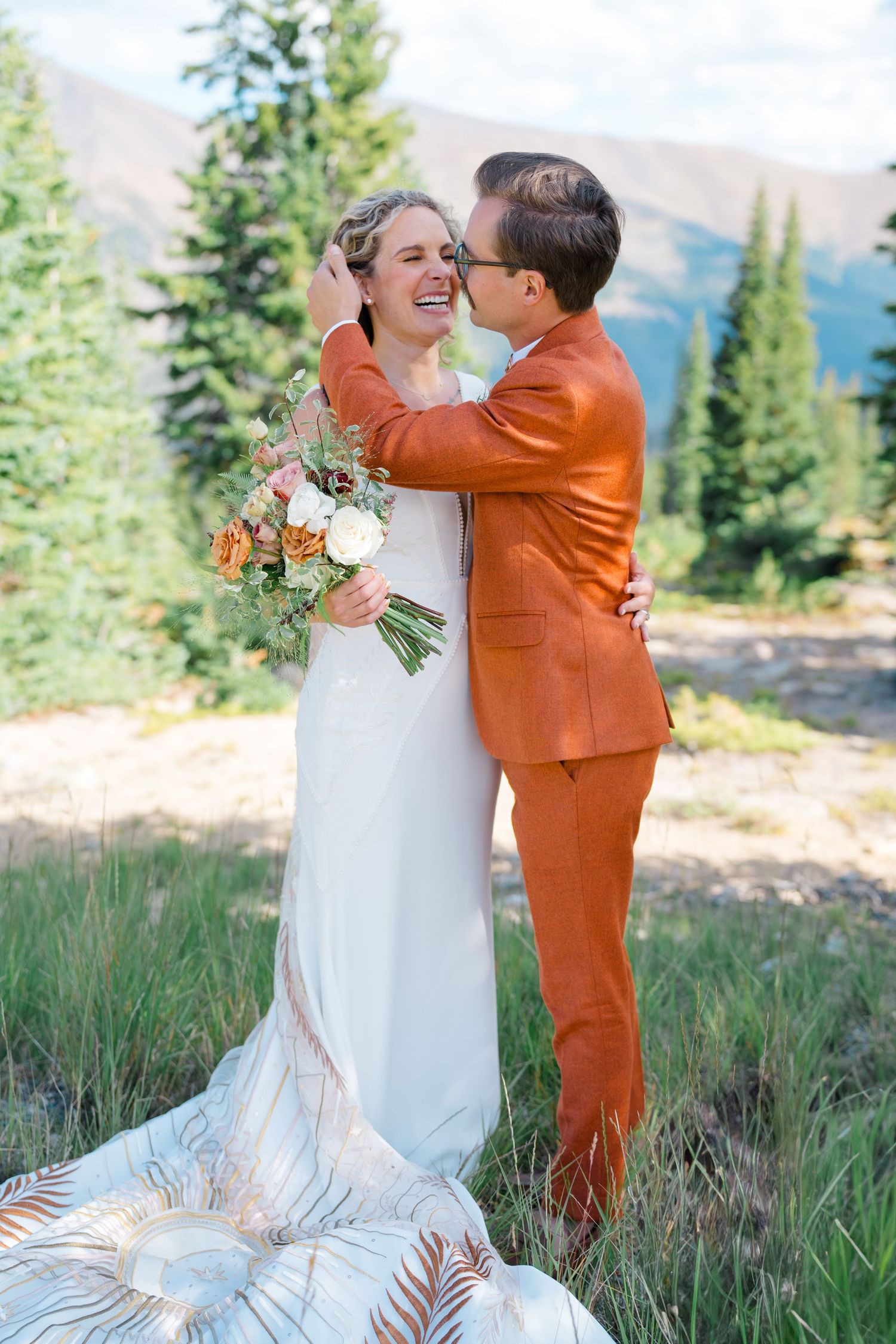 Wedding couple embracing in a mountain meadow with the bride in white and groom in vibrant orange suit captured by Vail Wedding Photographer