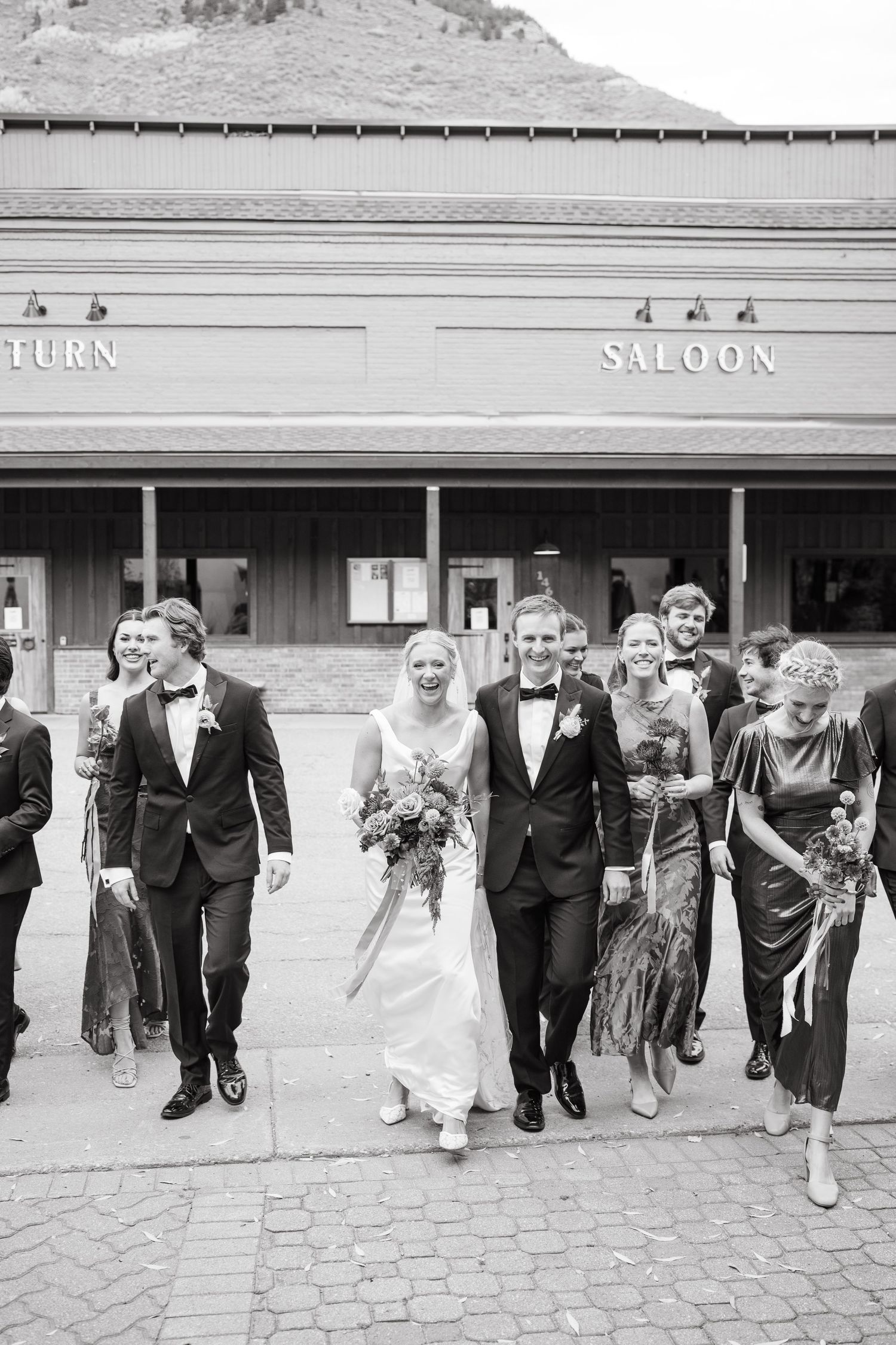 A wedding party walks together in front of a rustic saloon building.