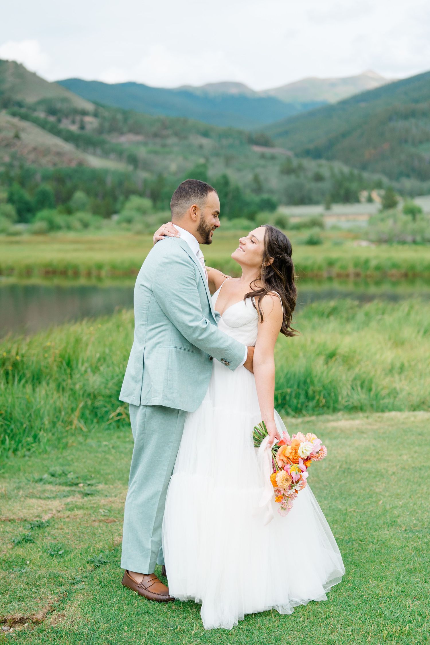 A couple shares a romantic moment in a white dress and mint suit against a scenic mountain landscape with rolling hills.
