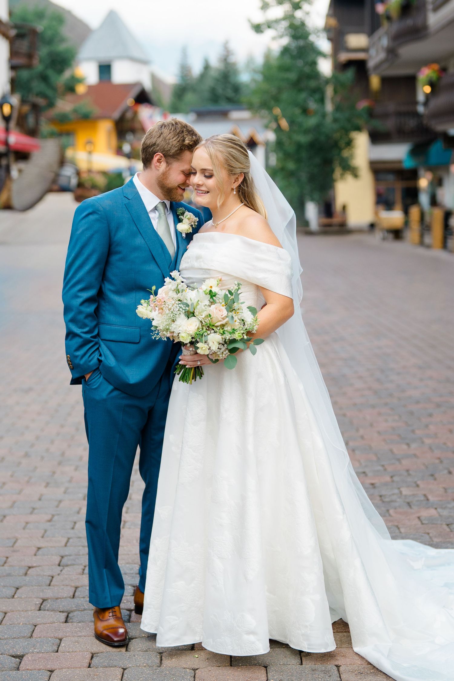 A couple in wedding attire shares intimate moments on a cobblestone street with mountains in the background.