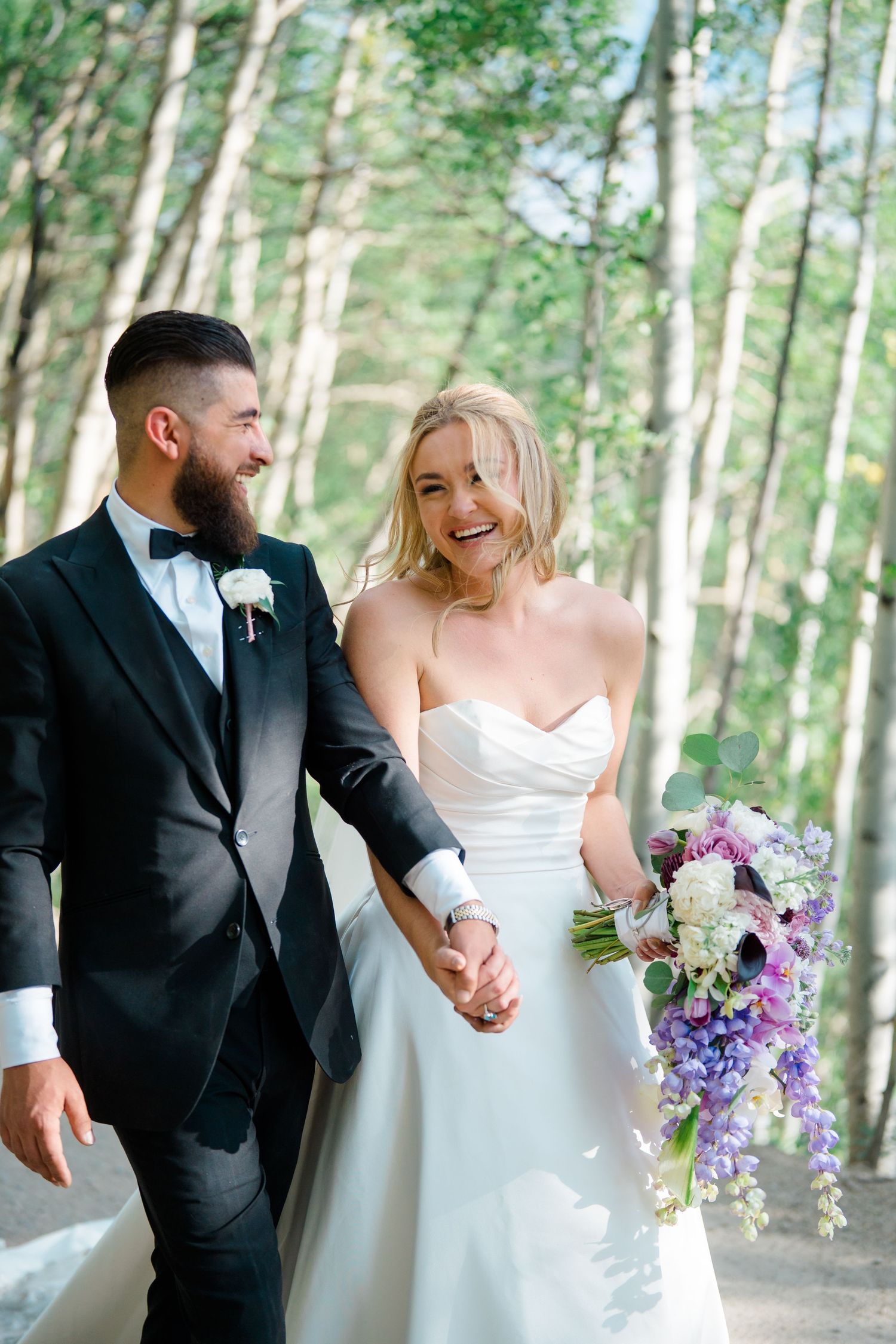 A couple shares joyful moments in a forest setting with purple and white wedding bouquet details.