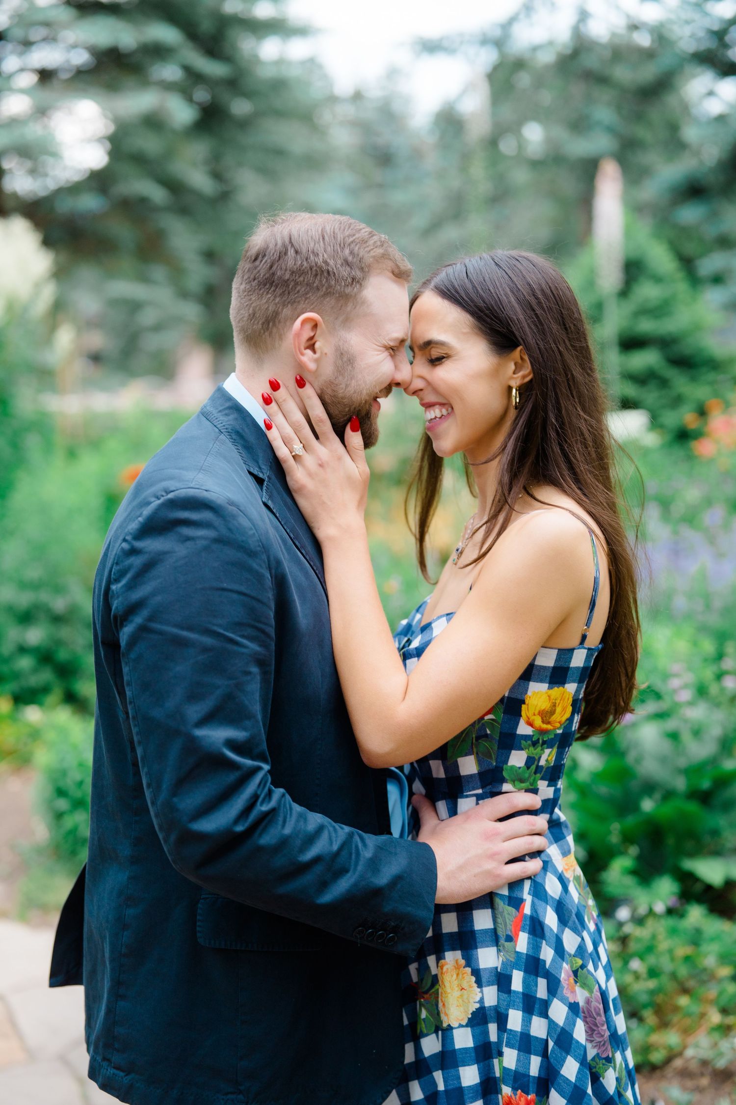 A couple shares a tender moment in a garden filled with evergreen trees and summer flowers.