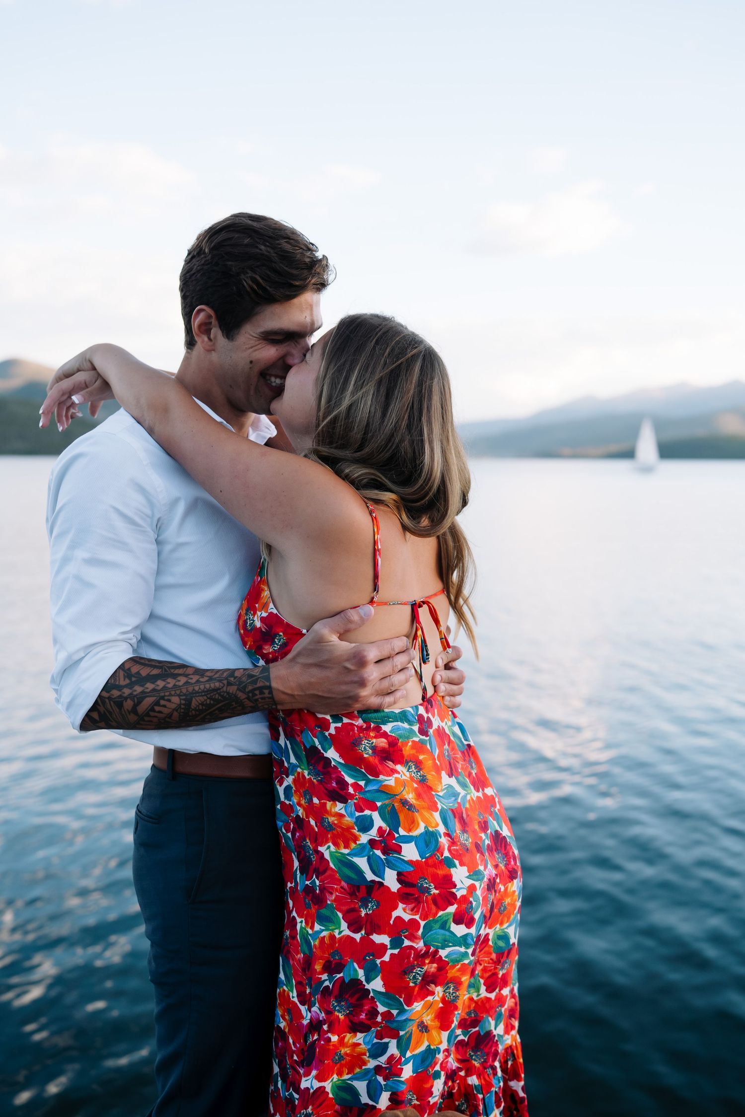 A couple embraces by the water at sunset with a sailboat visible in the distance.
