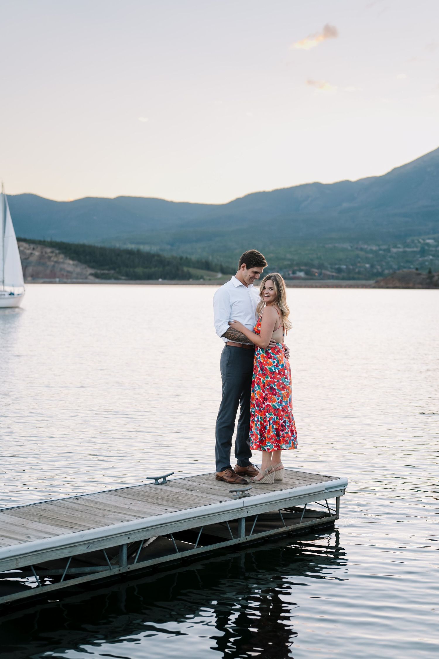 A couple stands together on a wooden dock overlooking a serene mountain lake with sailboats in the distance.
