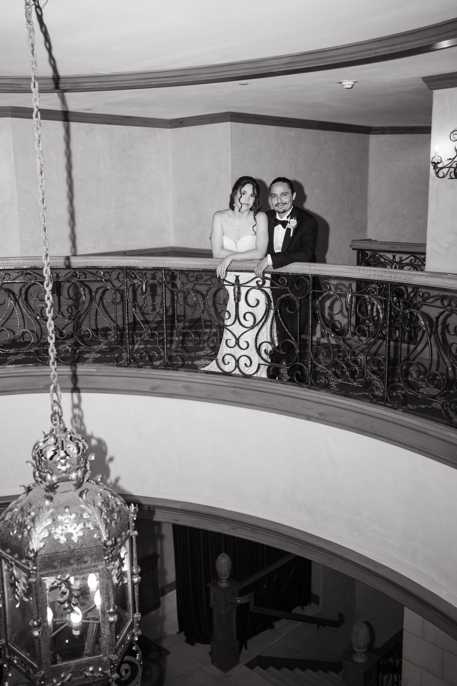 A couple poses on an elegant curved balcony with ornate iron railings and hanging pendant lights in a formal setting.