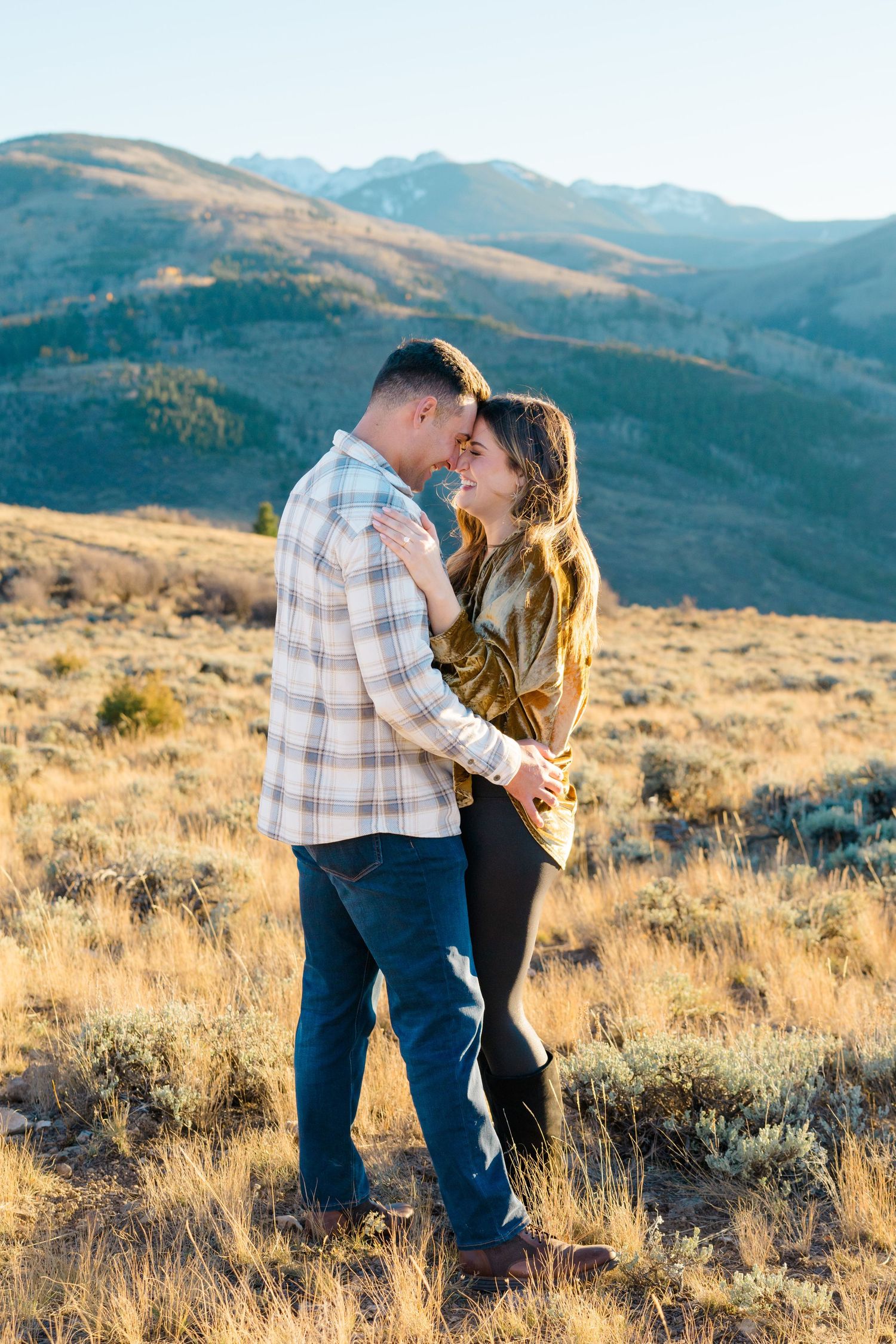 A couple embraces in a golden mountain meadow at sunset with rolling hills and sagebrush in the background.