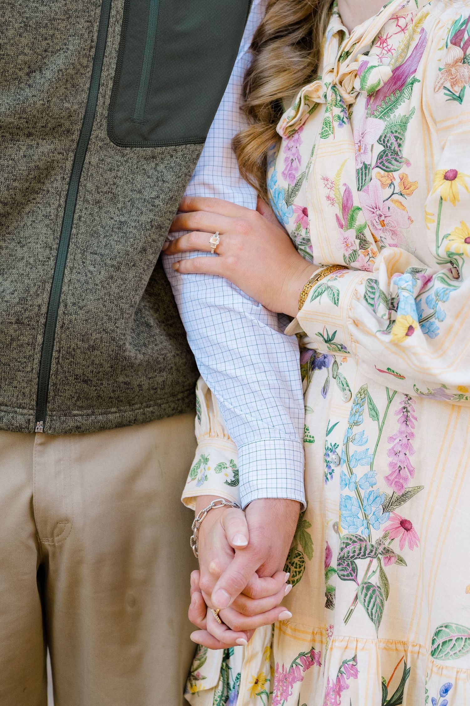 Couple holding hands, woman in floral dress with gold ring and bracelet, man in checkered shirt and khaki pants.