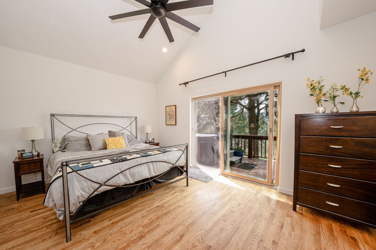 Bright bedroom with vaulted ceiling, hardwood floors, metal bed frame, dresser, ceiling fan, and sliding glass door to balcony.