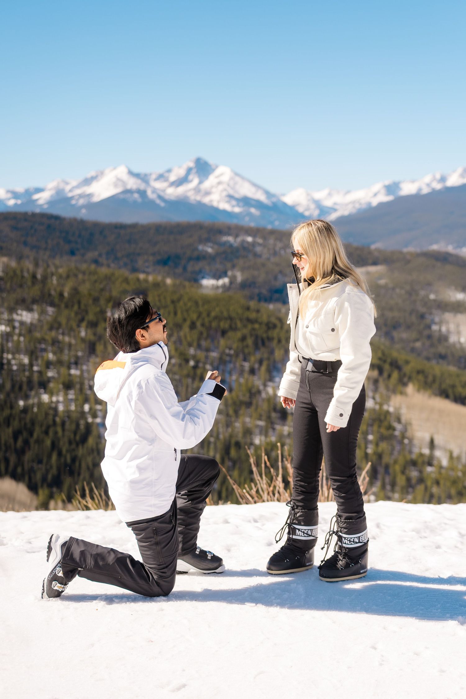 Man proposing on one knee to woman on snowy mountain top with scenic snow-capped peaks in background.