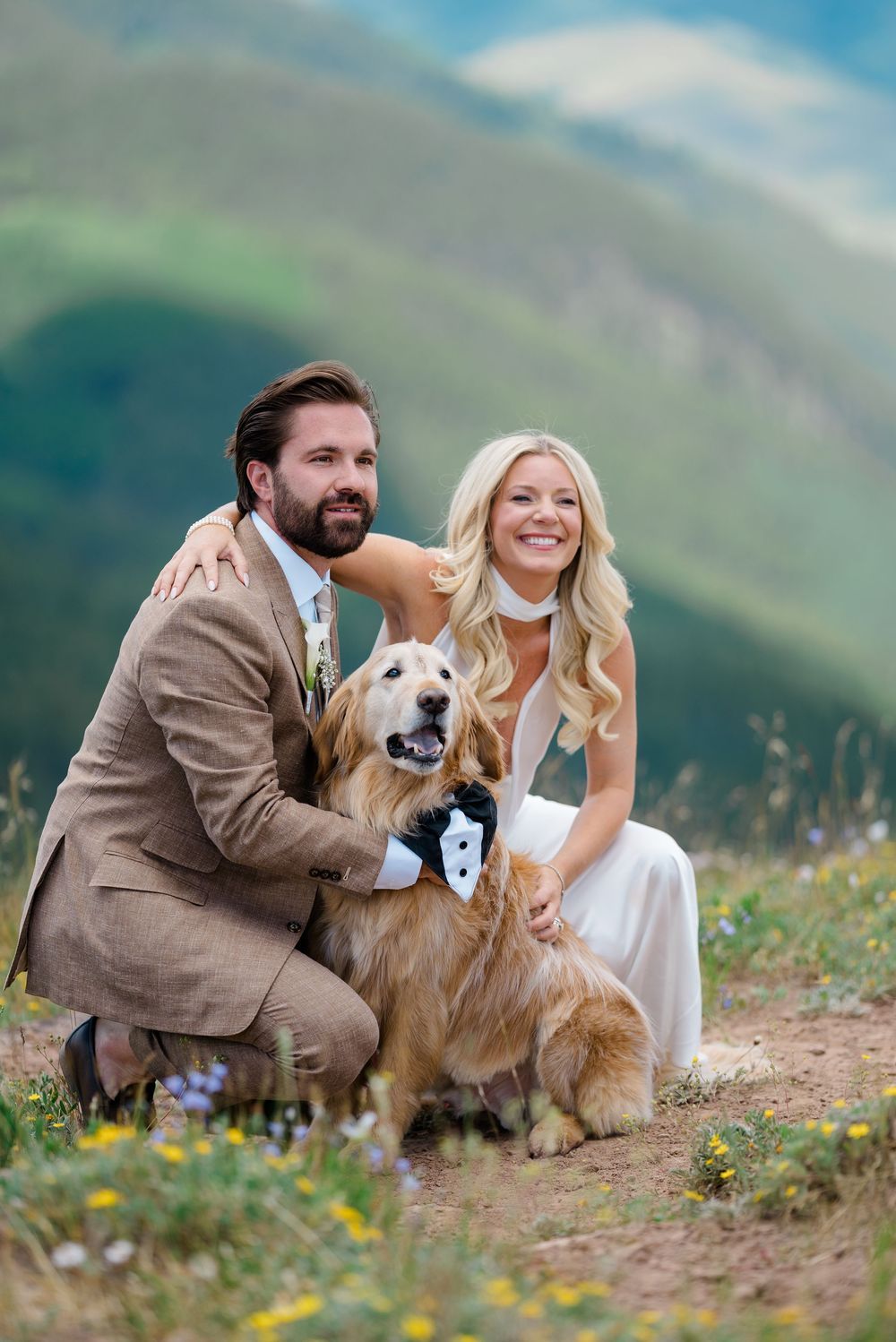 Bride and groom posing with their golden retriever in a tuxedo bandana on a scenic mountain meadow.